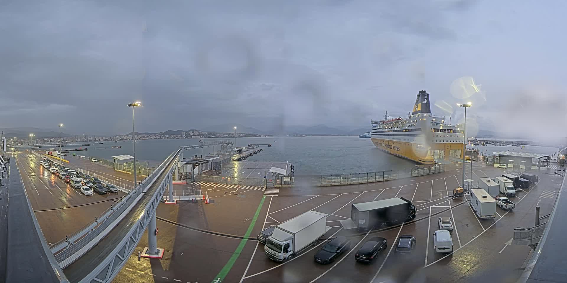 A large ferry is docked at a port on a cloudy, wet day, with numerous cars and trucks parked nearby.
