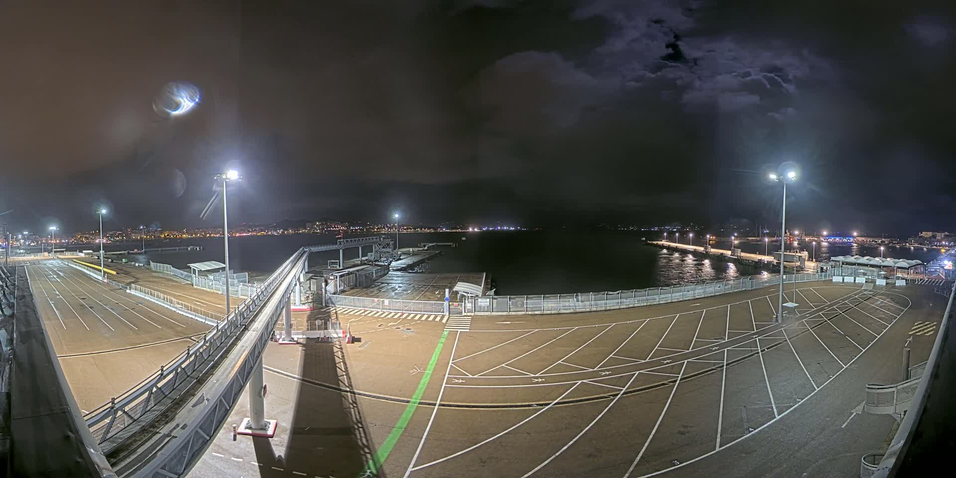 An empty, nighttime ferry terminal with parking areas and a gangway, overlooks a dark, choppy body of water under a cloudy sky.
