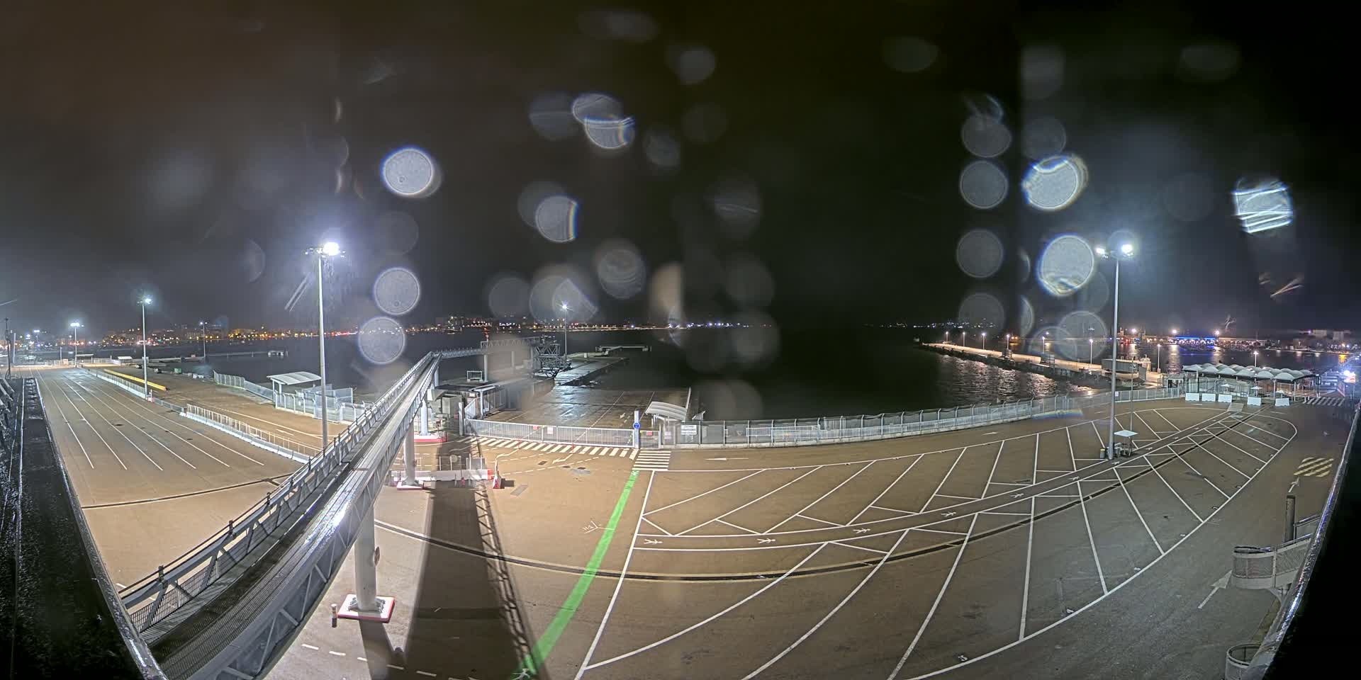 A nighttime view of a rain-slicked port area with empty parking lots, a gangway, and a dark body of water beyond, all illuminated by artificial lights.