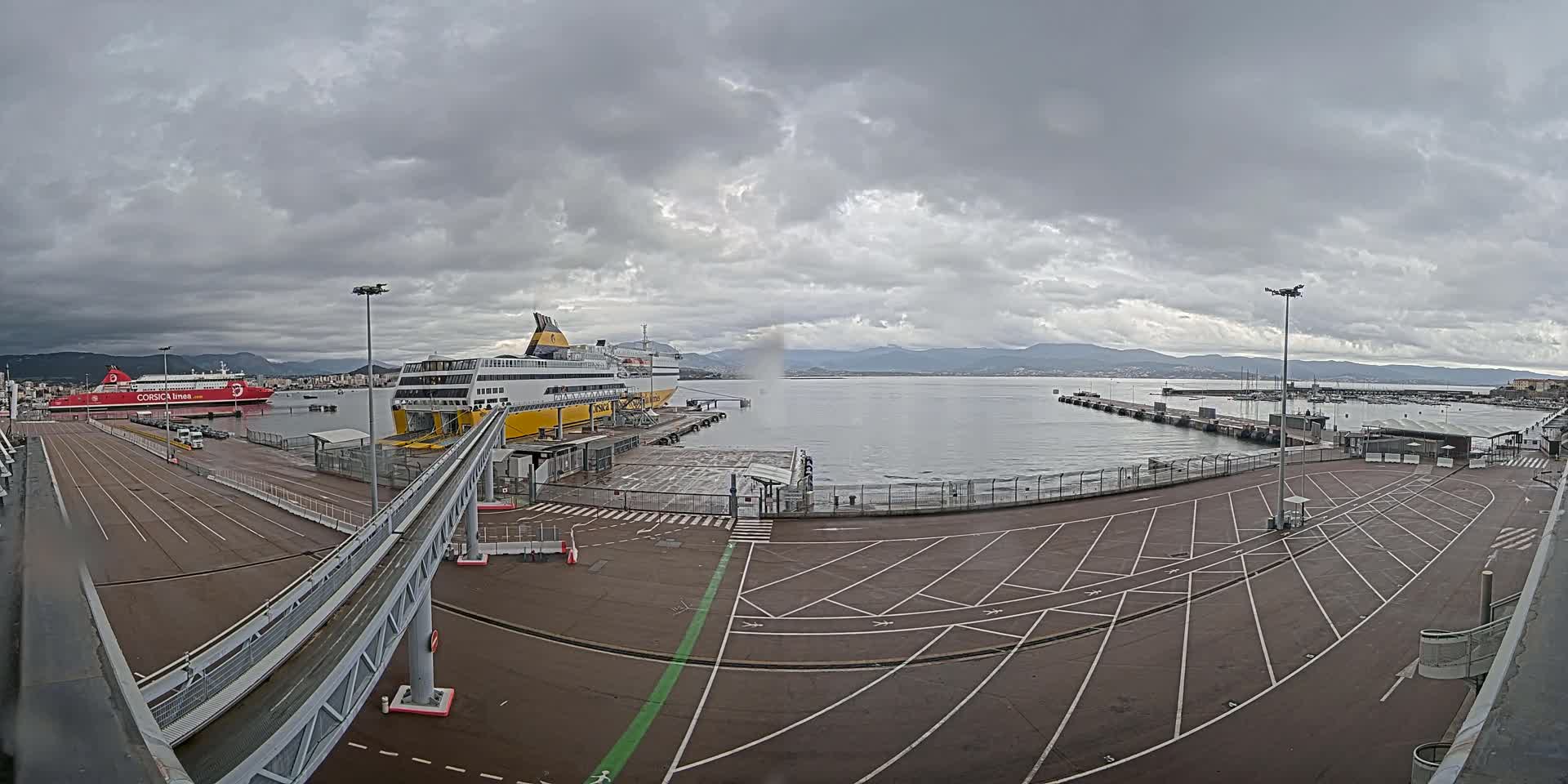 A ferry terminal with two docked ferries under a cloudy sky, overlooking a calm body of water and distant mountains.