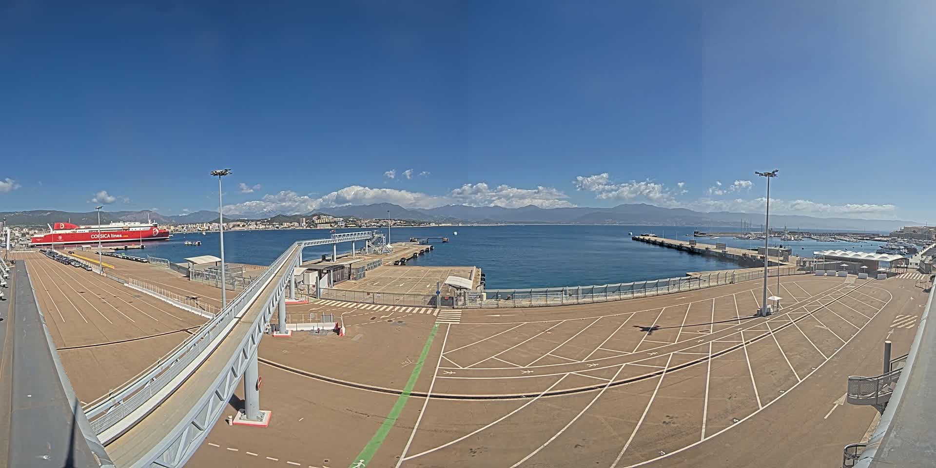 A large ferry is docked at a port on a sunny day with partly cloudy skies and mountains visible in the distance.
