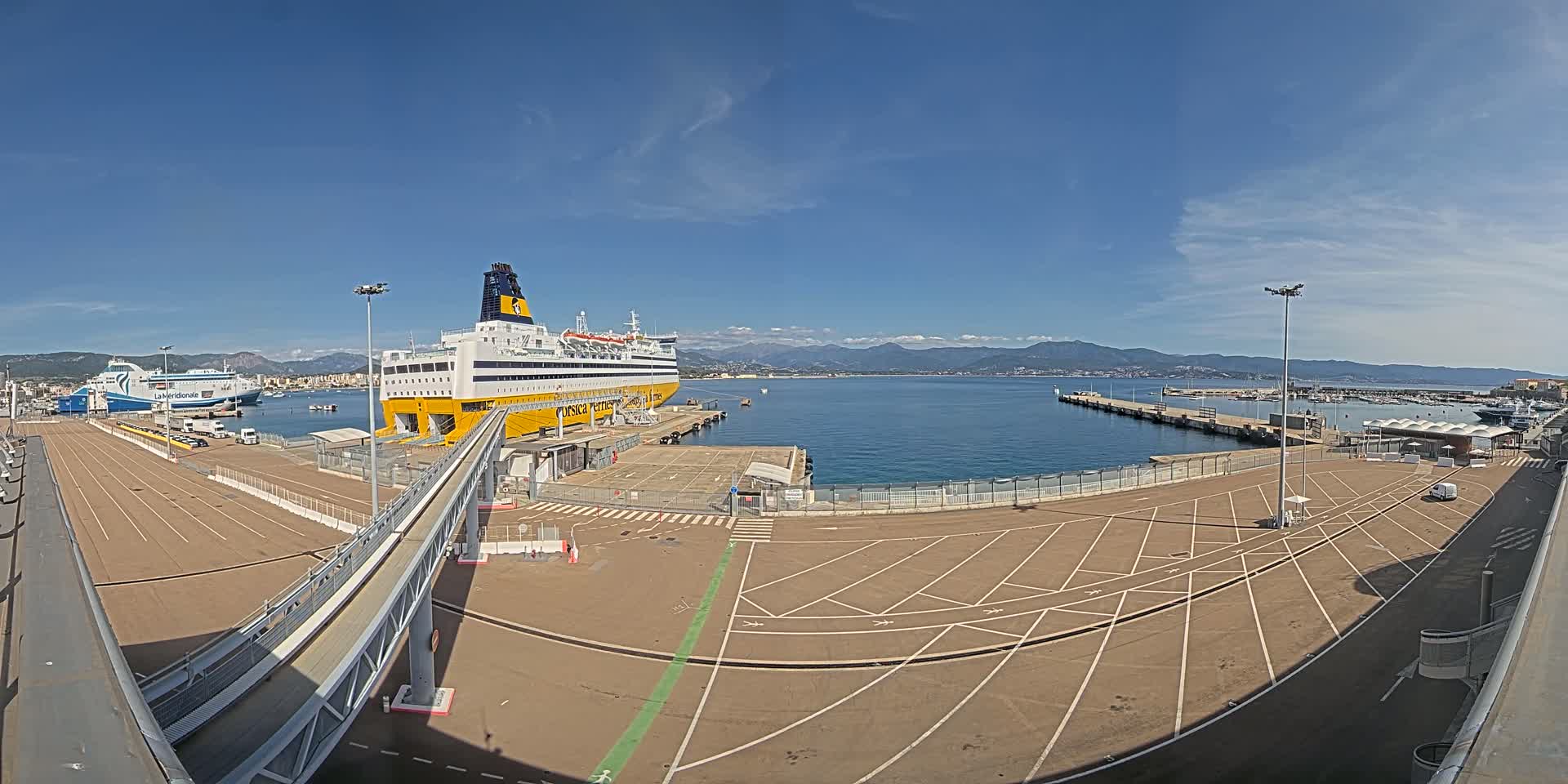 A large yellow and white ferry is docked at a port on a sunny day, with mountains visible in the background and another smaller ferry further down the dock.