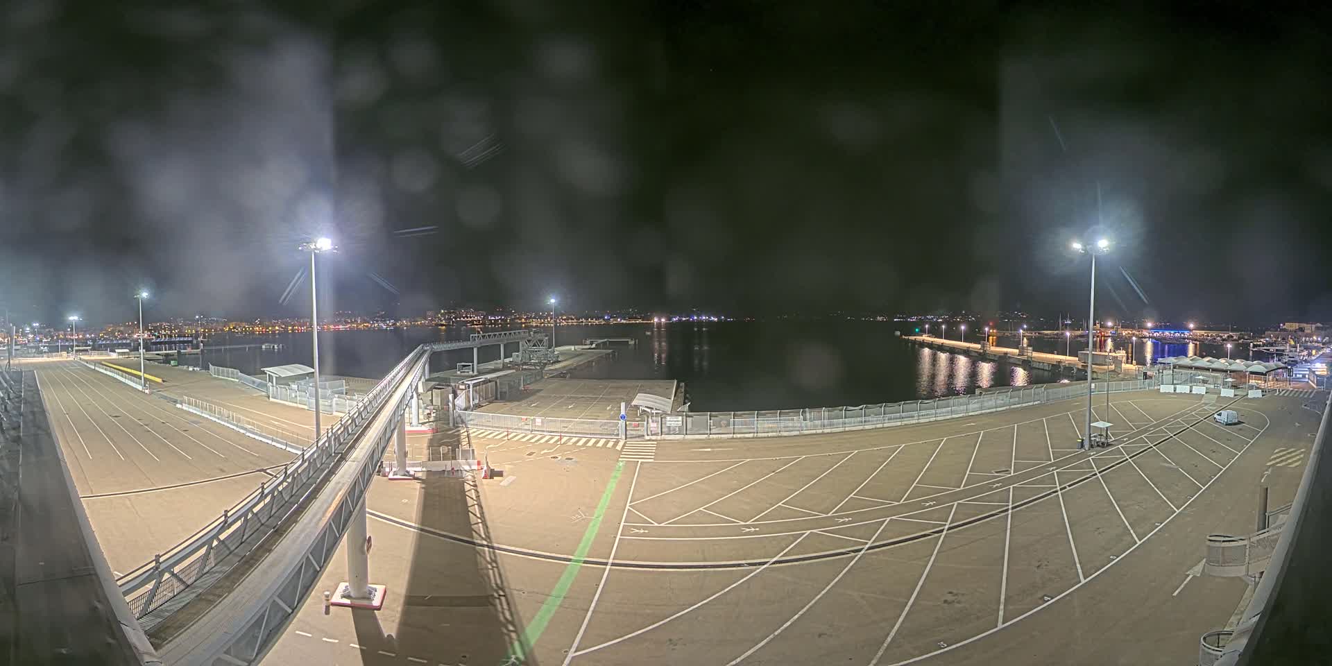 An empty, well-lit port at night under a slightly hazy sky, with a walkway leading to a dock extending into calm water.