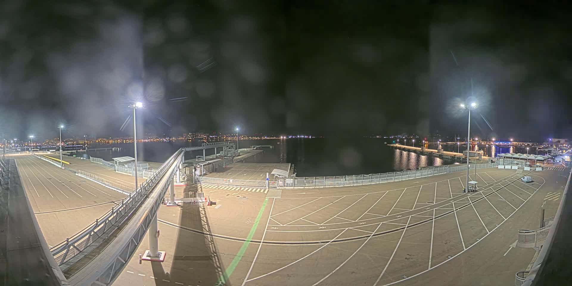 A nighttime view of a mostly empty port area with a gangway, parking lot, and body of water beyond a fence, under slightly hazy conditions.