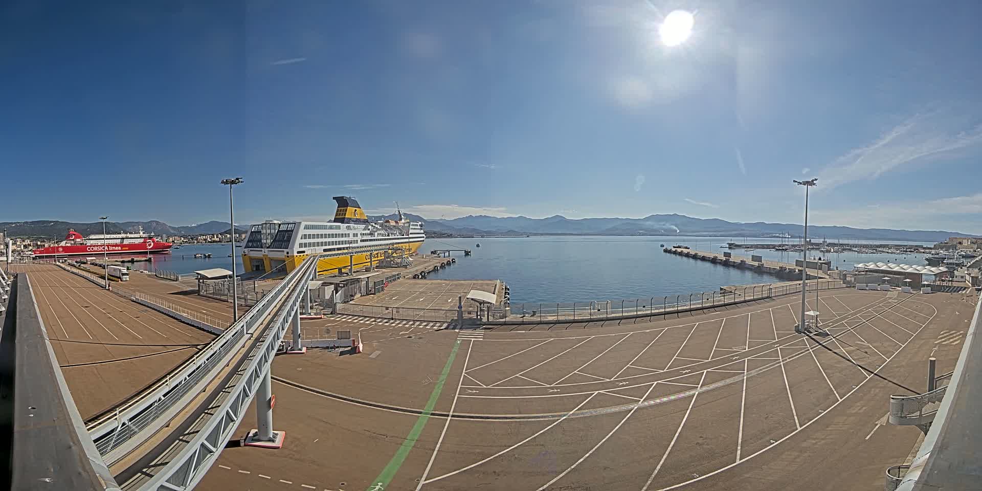 A sunny day at a port shows a large yellow and black ferry docked, a smaller red ferry in the background, a paved area with parking spaces, and mountains visible across the calm water.