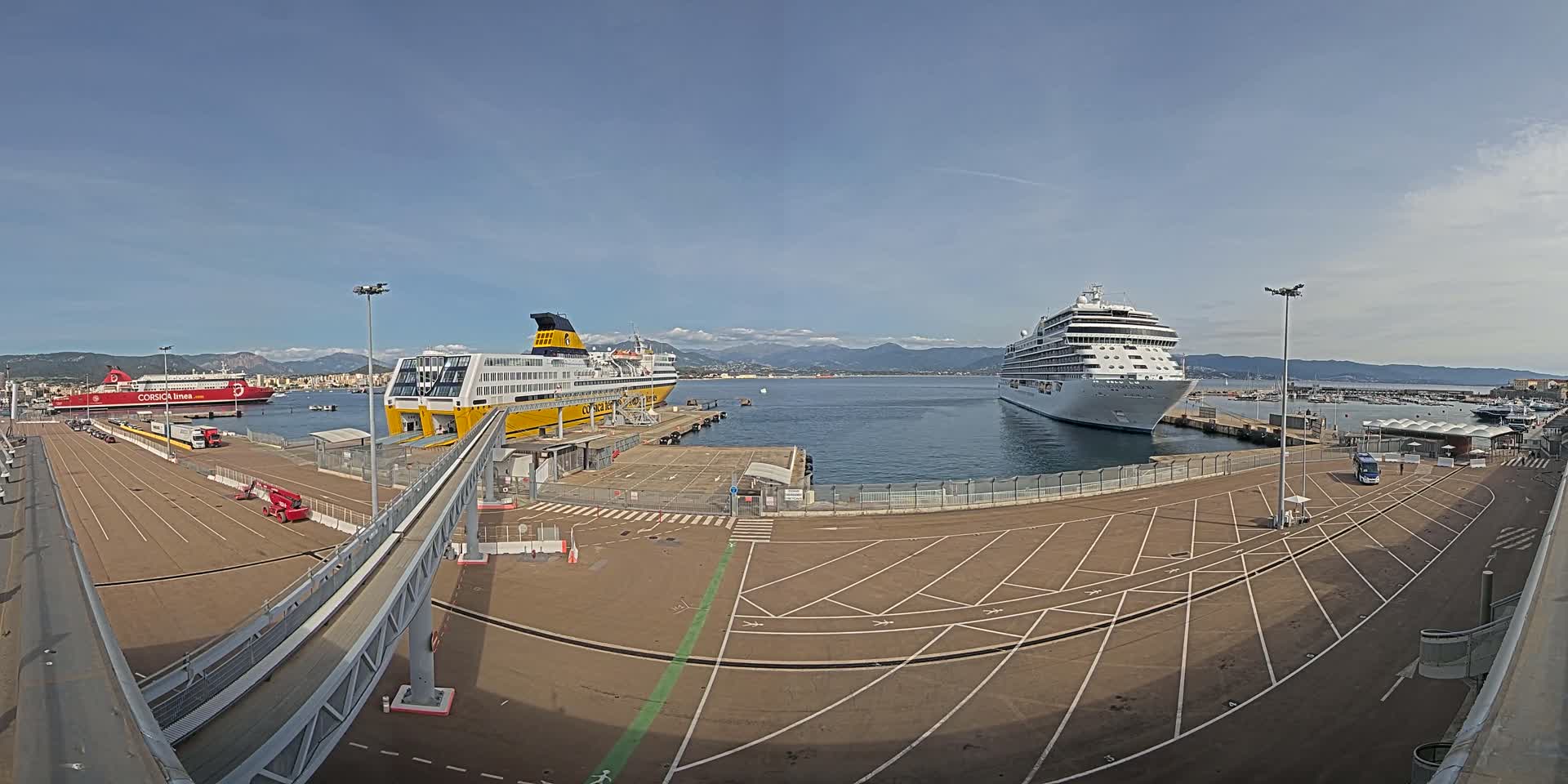 A large cruise ship and two ferries are docked at a port on a mostly sunny day with mountains in the background.