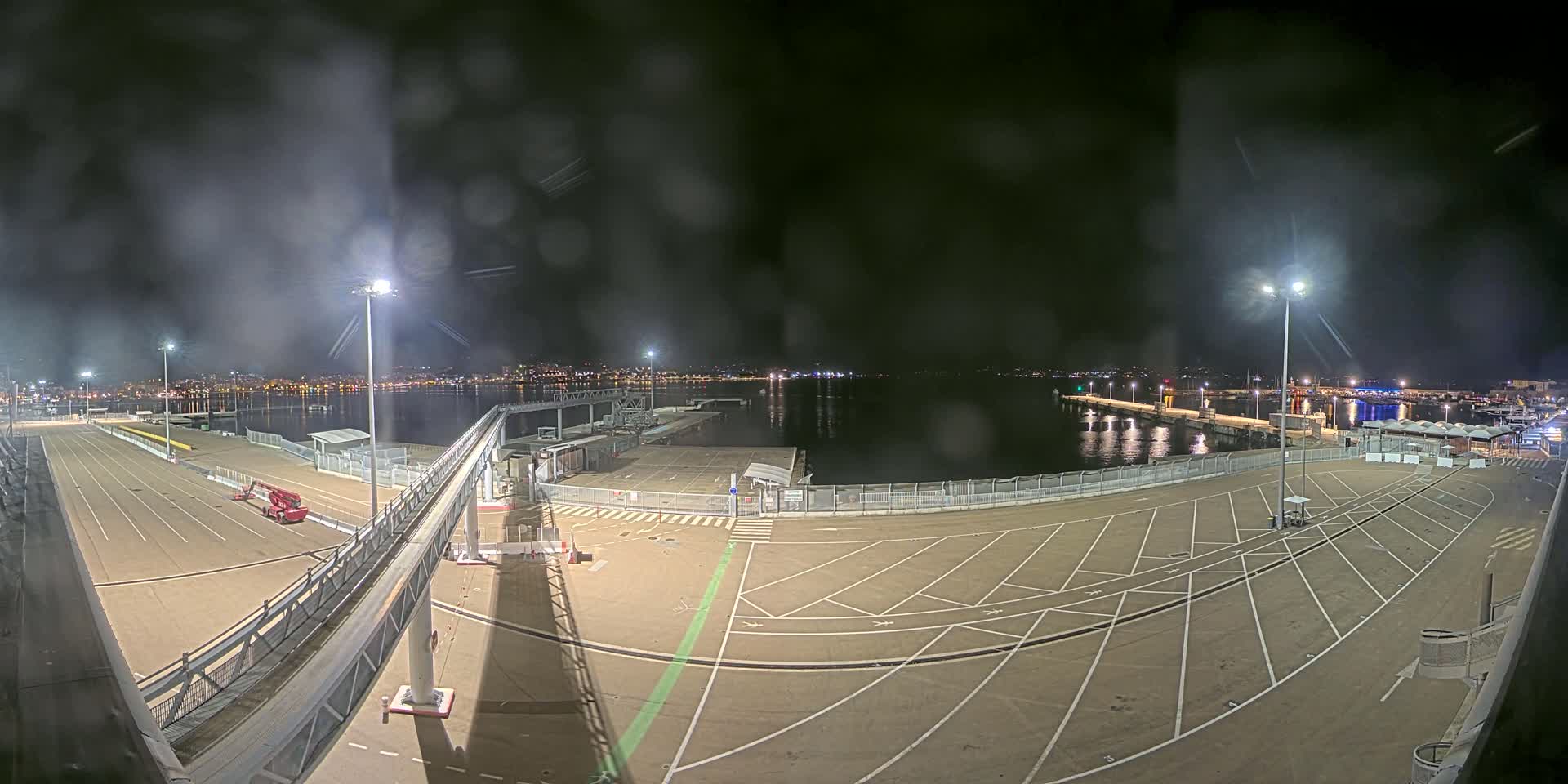 A nighttime view of a port with empty parking lots, a gangway, and a calm body of water reflecting city lights in the distance under a slightly hazy sky.
