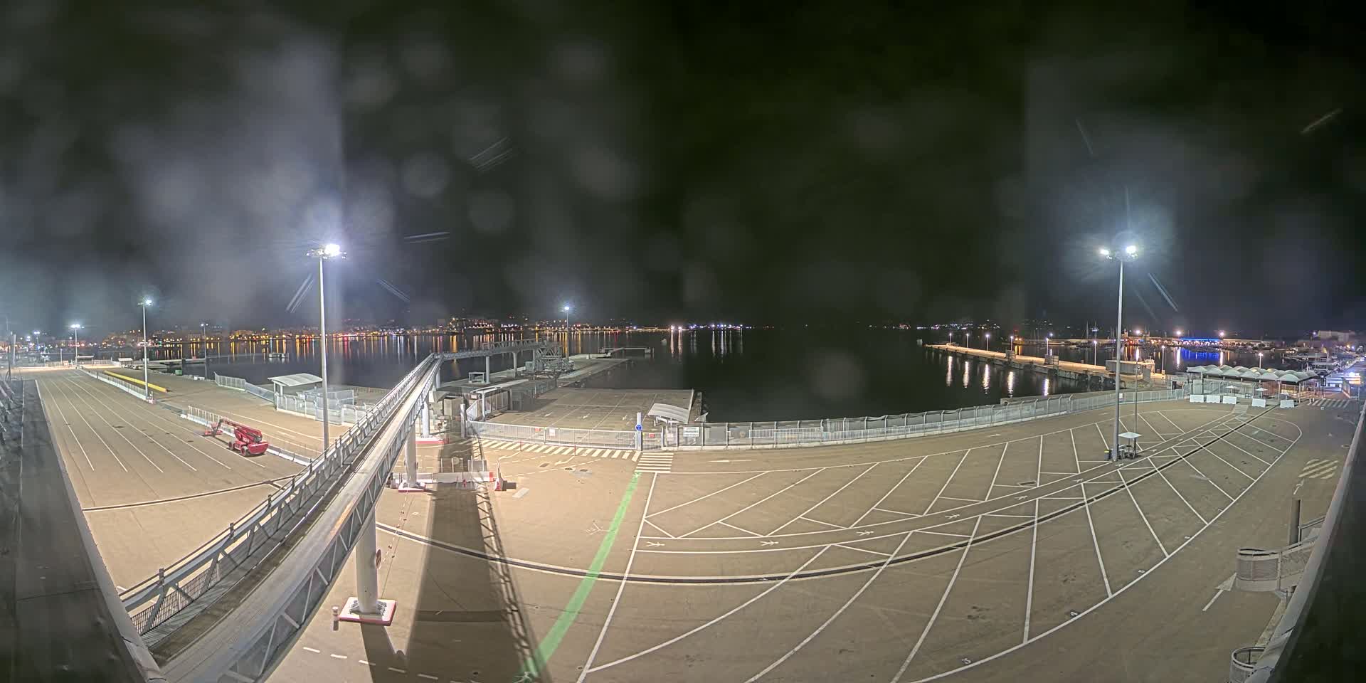 A nighttime view of an empty port parking lot and a calm body of water with lights visible across the water and on the far shore, under hazy conditions.