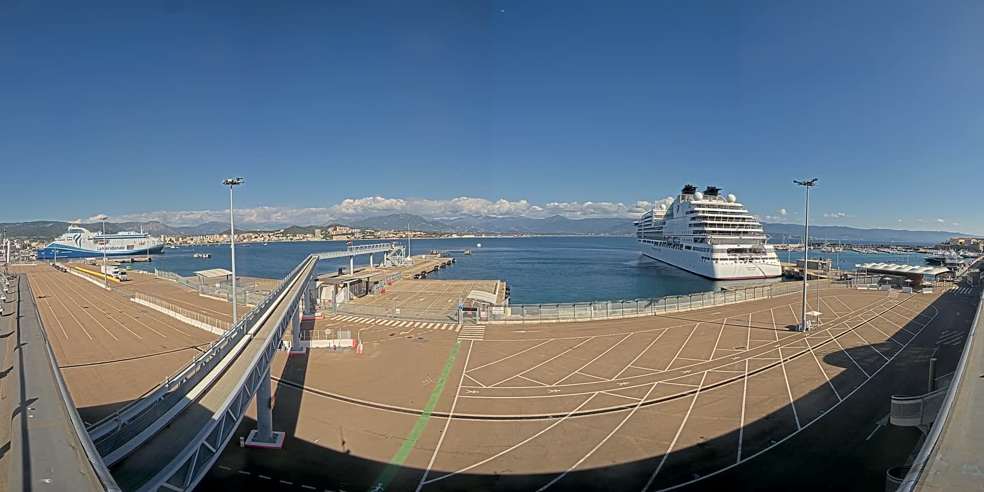 A large cruise ship and a ferry are docked at a port on a sunny day with mountains visible in the distance.