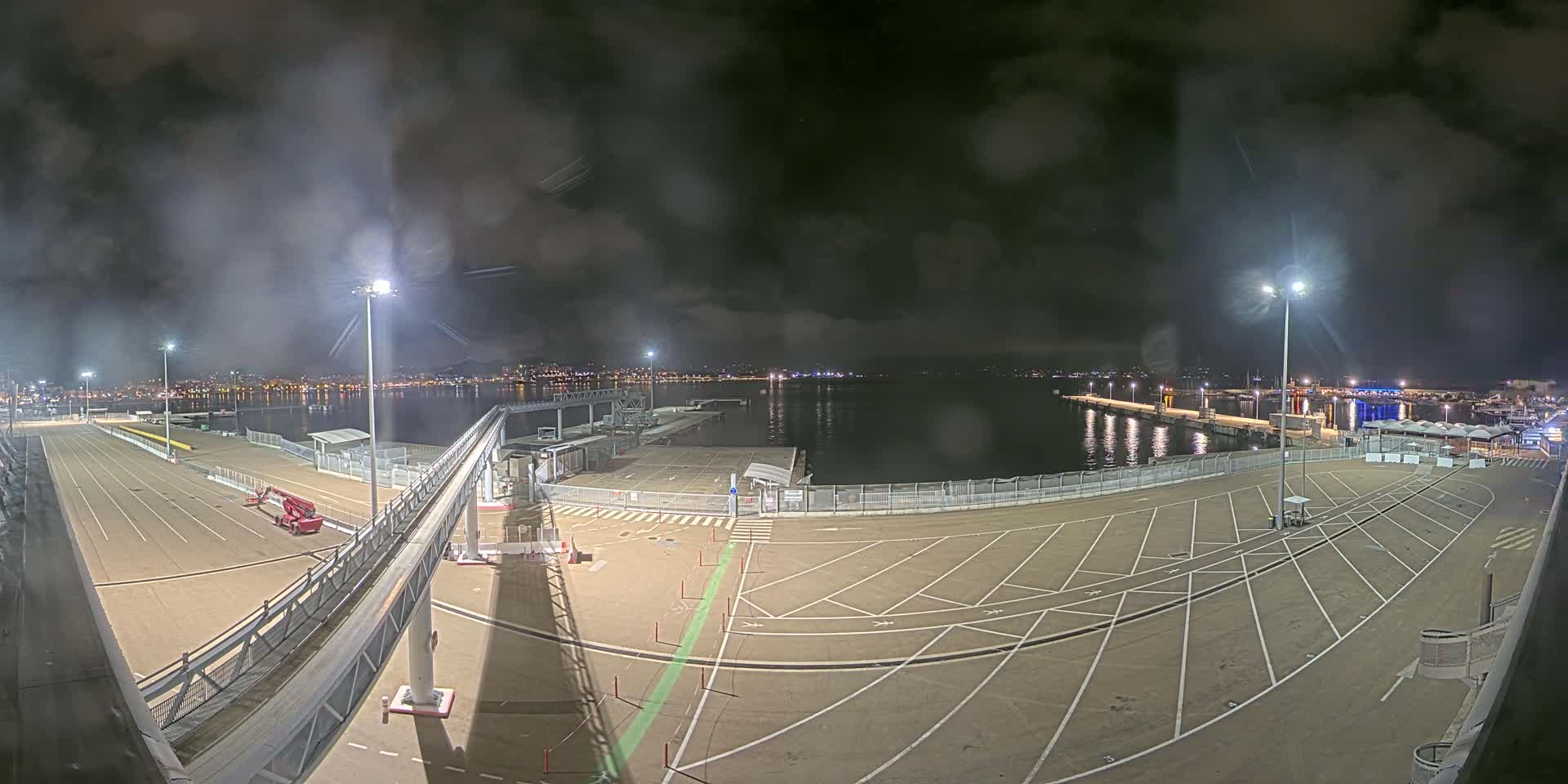 An empty, nighttime port area with a walkway and a city skyline visible across the water under a cloudy sky.