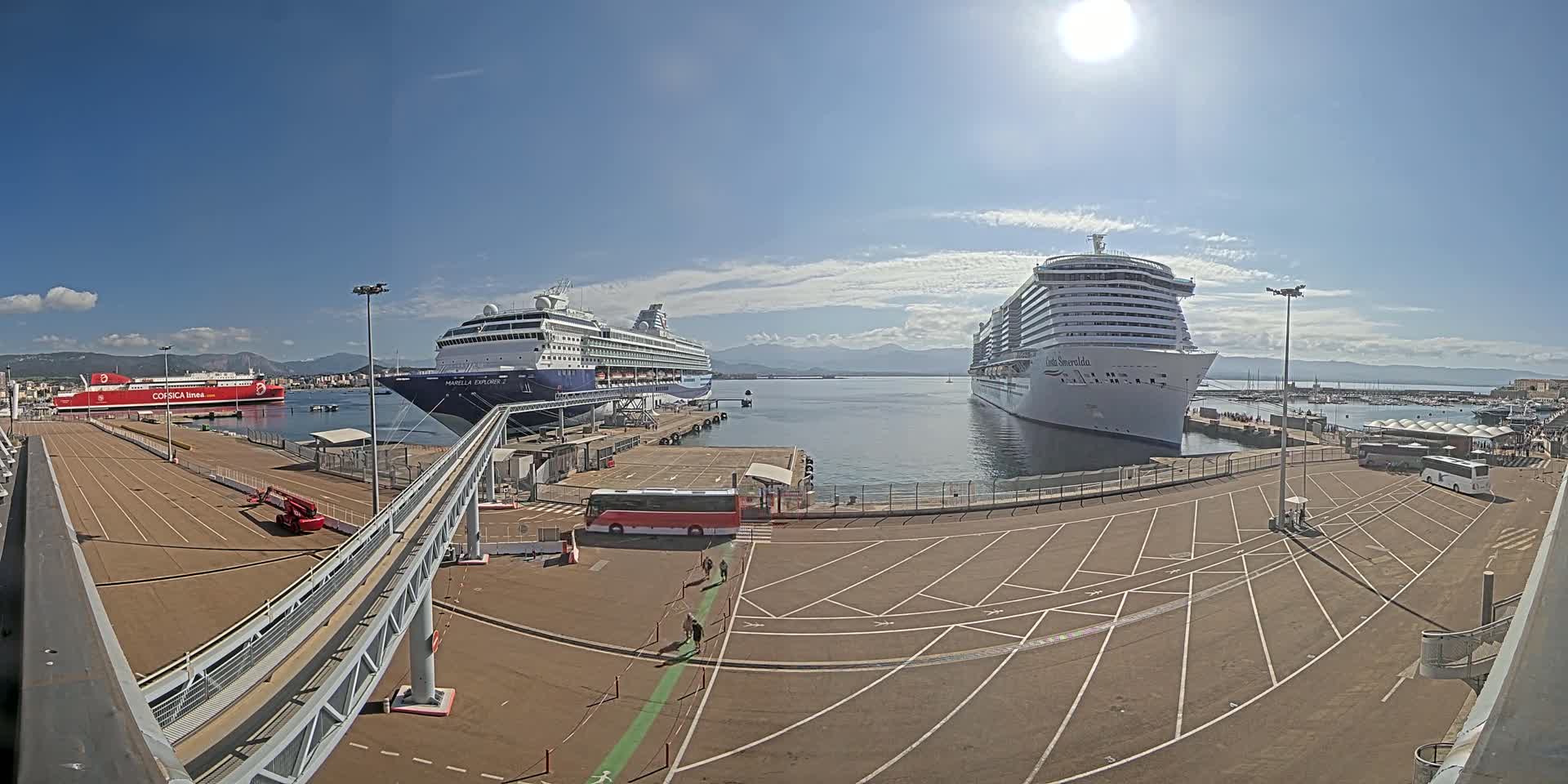 Two large cruise ships are docked at a port on a sunny day, with a ferry and mountains visible in the distance.