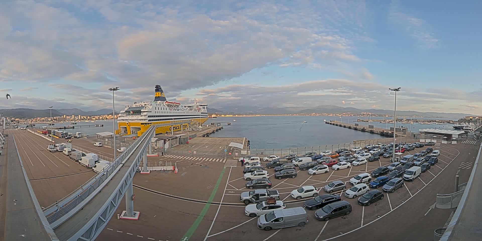 A large ferry is docked at a port under a partly cloudy sky, with numerous cars parked nearby waiting to board.