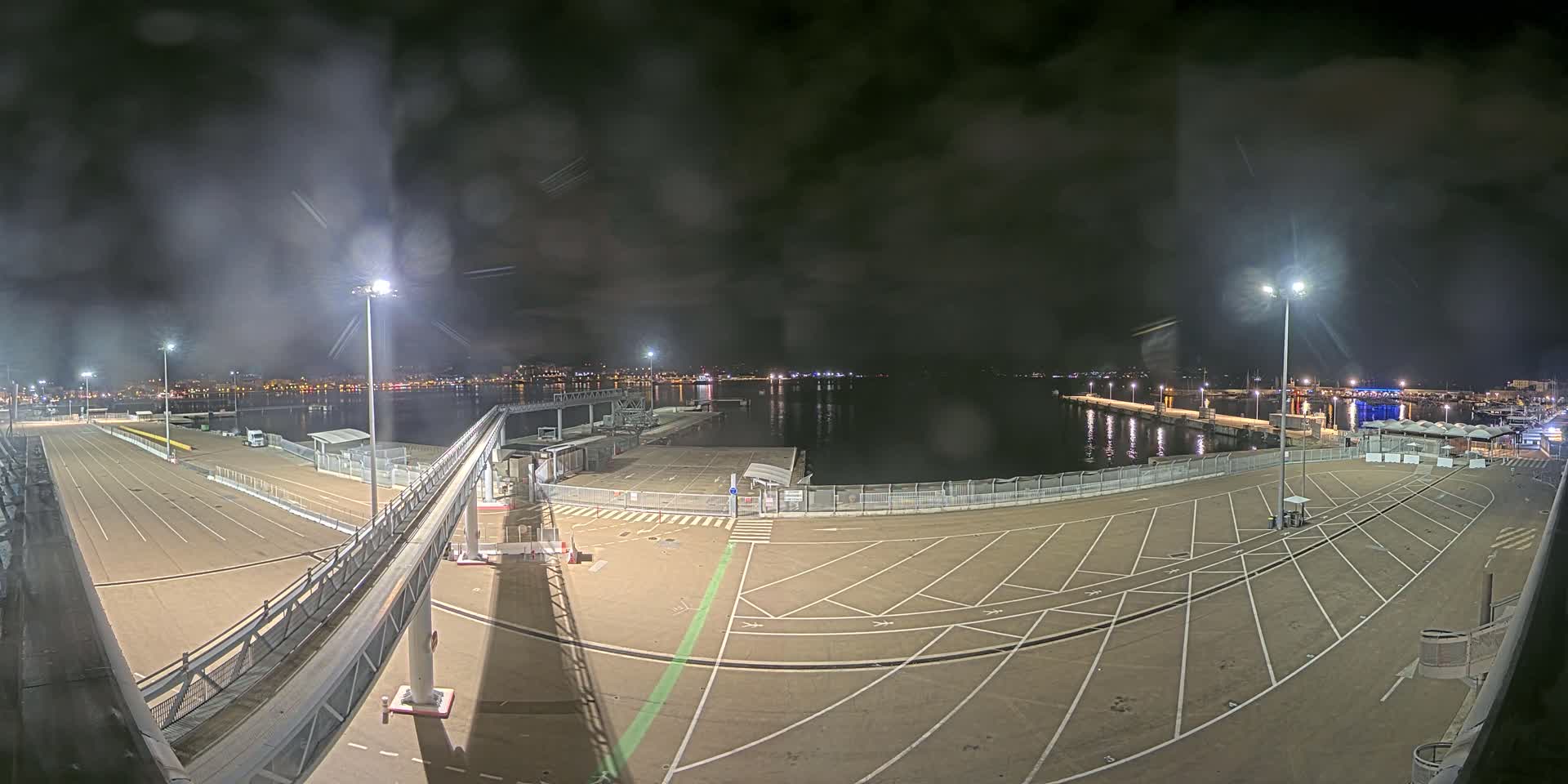 An empty, nighttime port parking lot with a gangway leading to a body of water, under a cloudy sky illuminated by artificial lights.