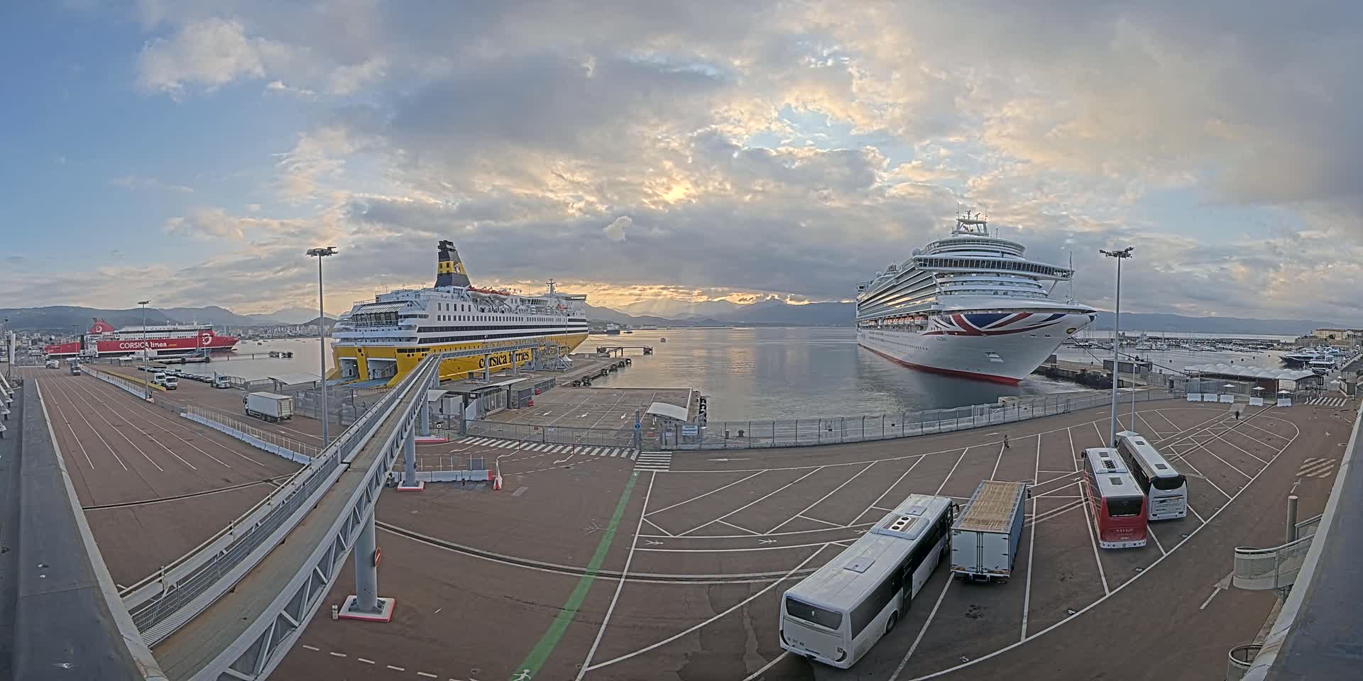 A large cruise ship and a ferry are docked at a port under a partly cloudy sky, with buses and other vehicles parked nearby.