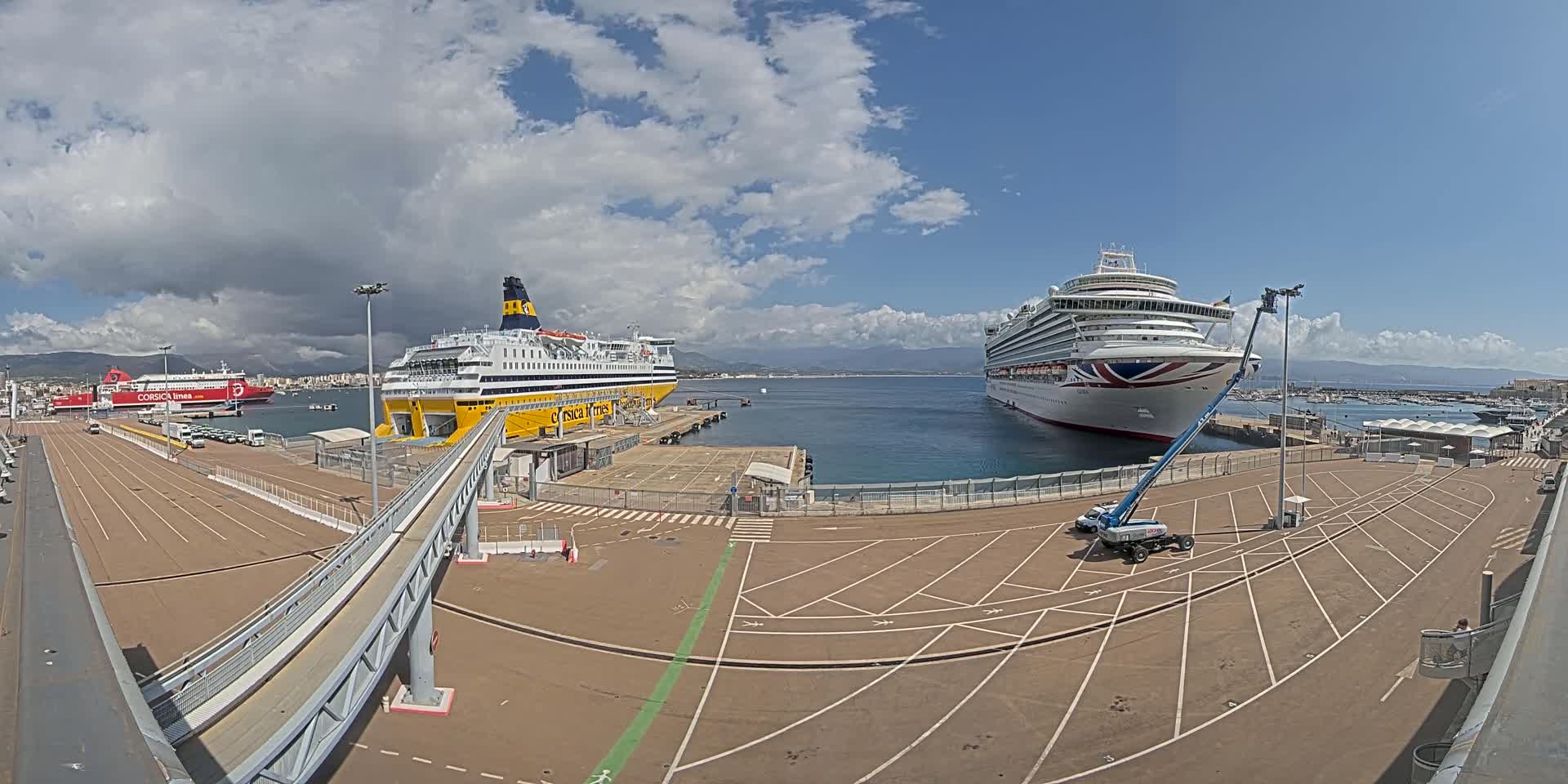 A large cruise ship and a ferry are docked at a port under a partly cloudy sky.