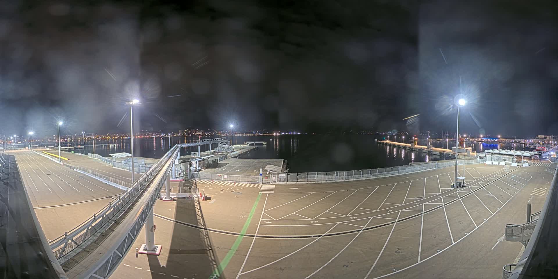An empty nighttime port with a walkway leading to a dock, illuminated by several tall lights under a cloudy sky.