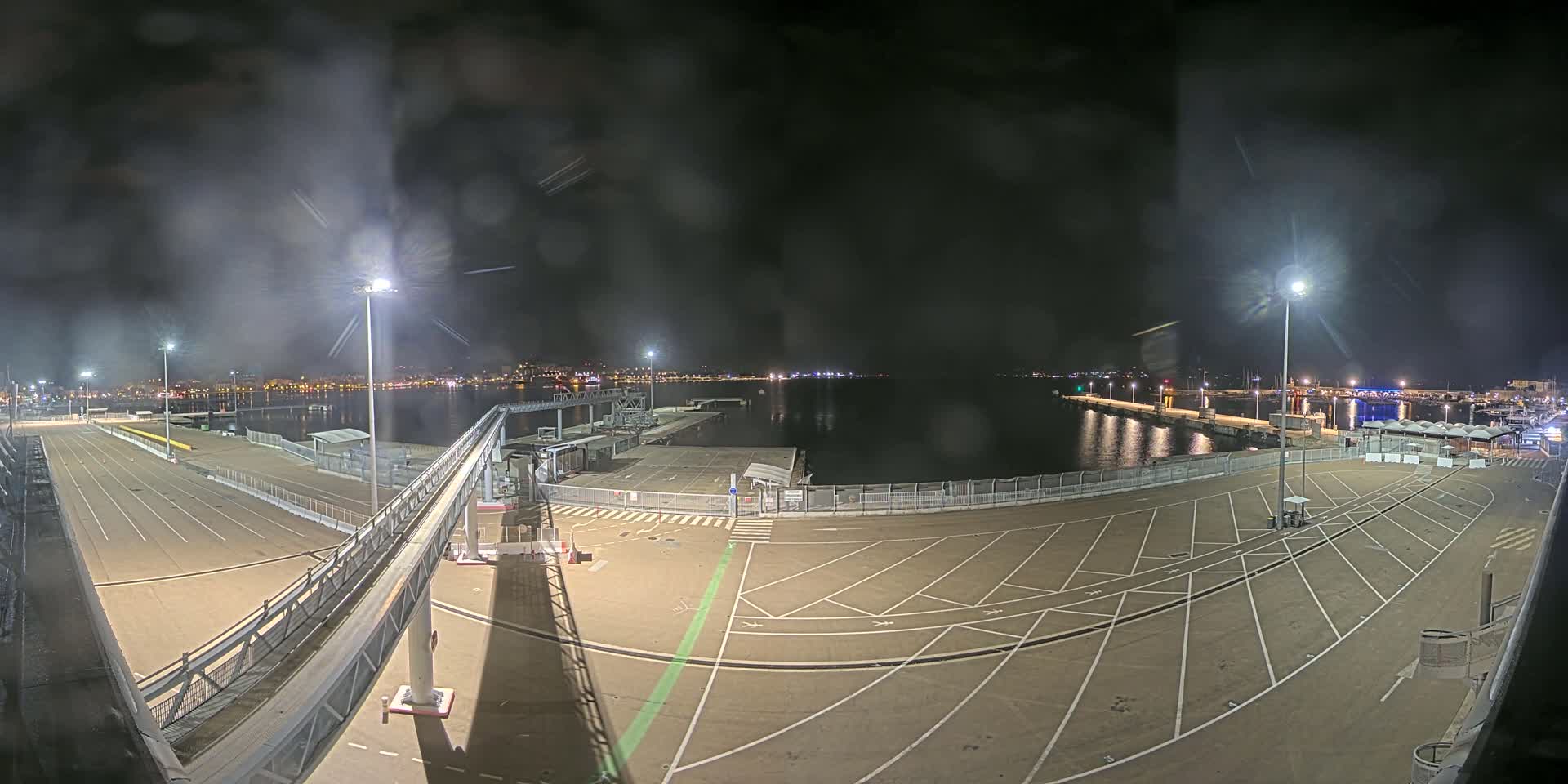 A nighttime view of an empty port area with a walkway leading to a dock, overlooking calm water and a distant city skyline under a dark, slightly hazy sky.
