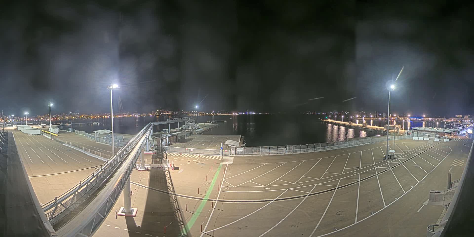 A nighttime view of an empty port area with a long gangway, illuminated by bright lights, overlooking a calm body of water and a city skyline in the distance.