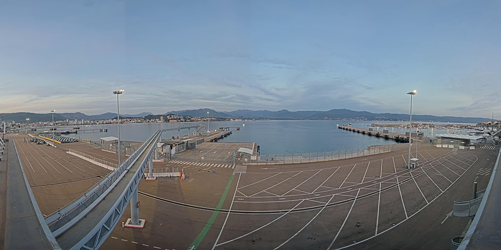 An empty parking lot at a ferry terminal overlooks a calm body of water with mountains in the distance under a clear twilight sky.