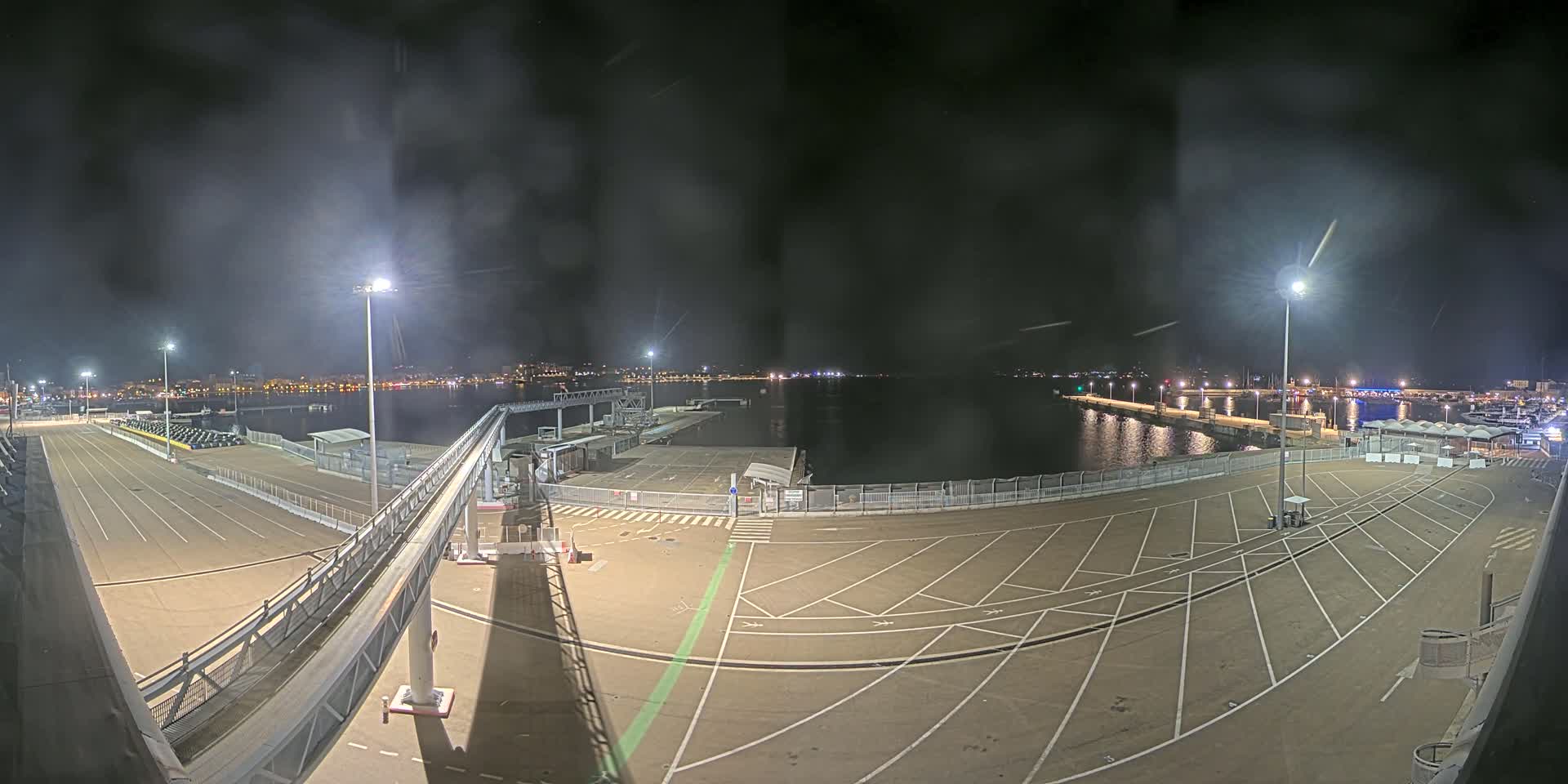 A nighttime view of an empty port area with a gangway leading to a dock, illuminated by bright lights, overlooking a calm body of water and a distant city skyline.