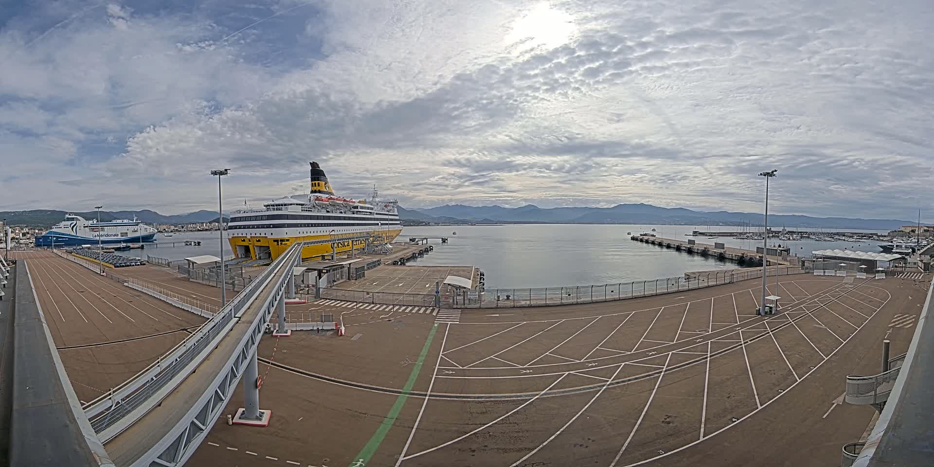 A large ferry is docked at a port under a partly cloudy sky, with another ferry and numerous boats visible in the distance.