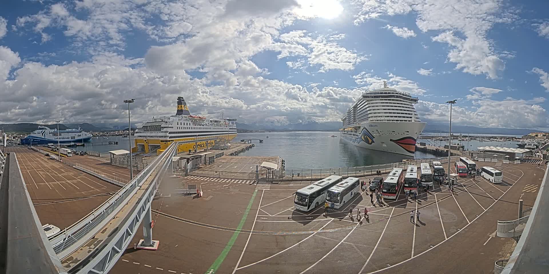 A sunny day at a harbor shows two large passenger ships docked, several buses parked nearby, and a walkway leading to one of the ships.