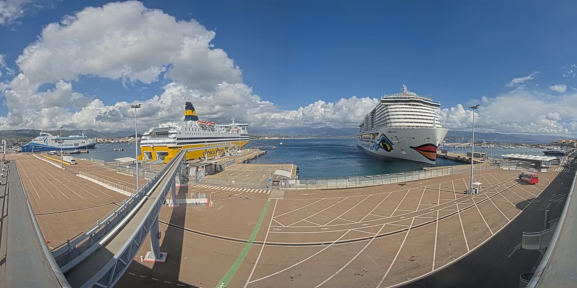 A large cruise ship and a ferry are docked at a port under a partly cloudy sky.