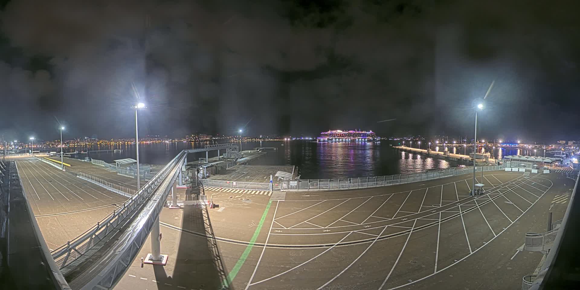 A nighttime view of an empty port area with a large cruise ship in the water under a cloudy sky.