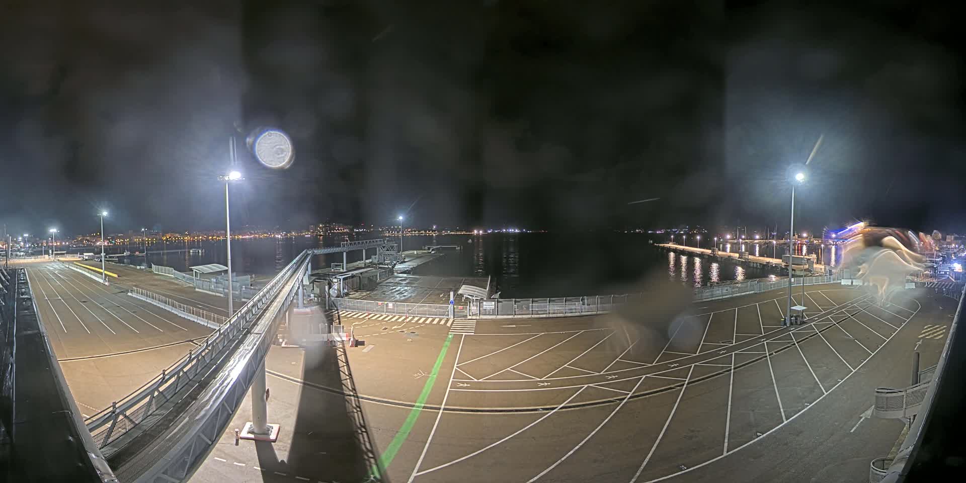 A nighttime view of an empty port with rain on the camera lens, showing a dark body of water, a city skyline in the distance, and marked parking areas.