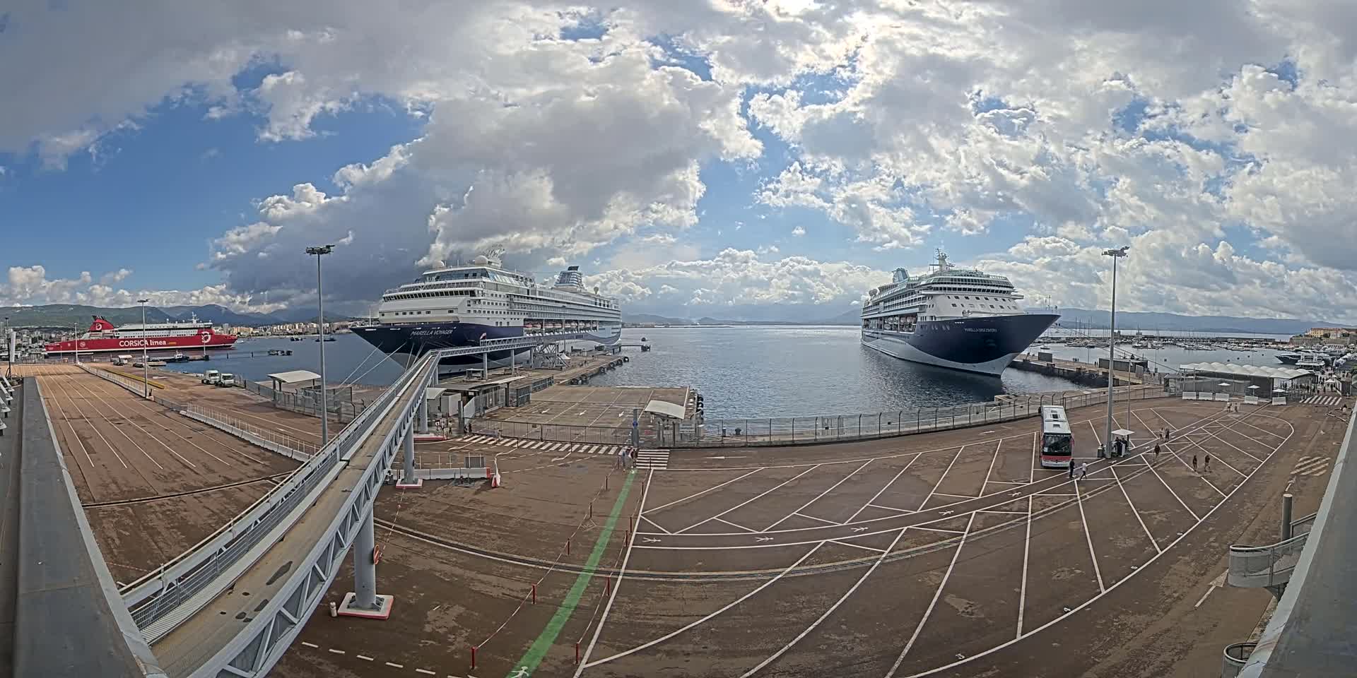 Two large cruise ships are docked at a port under a partly cloudy sky, with a ferry and smaller boats visible in the background.