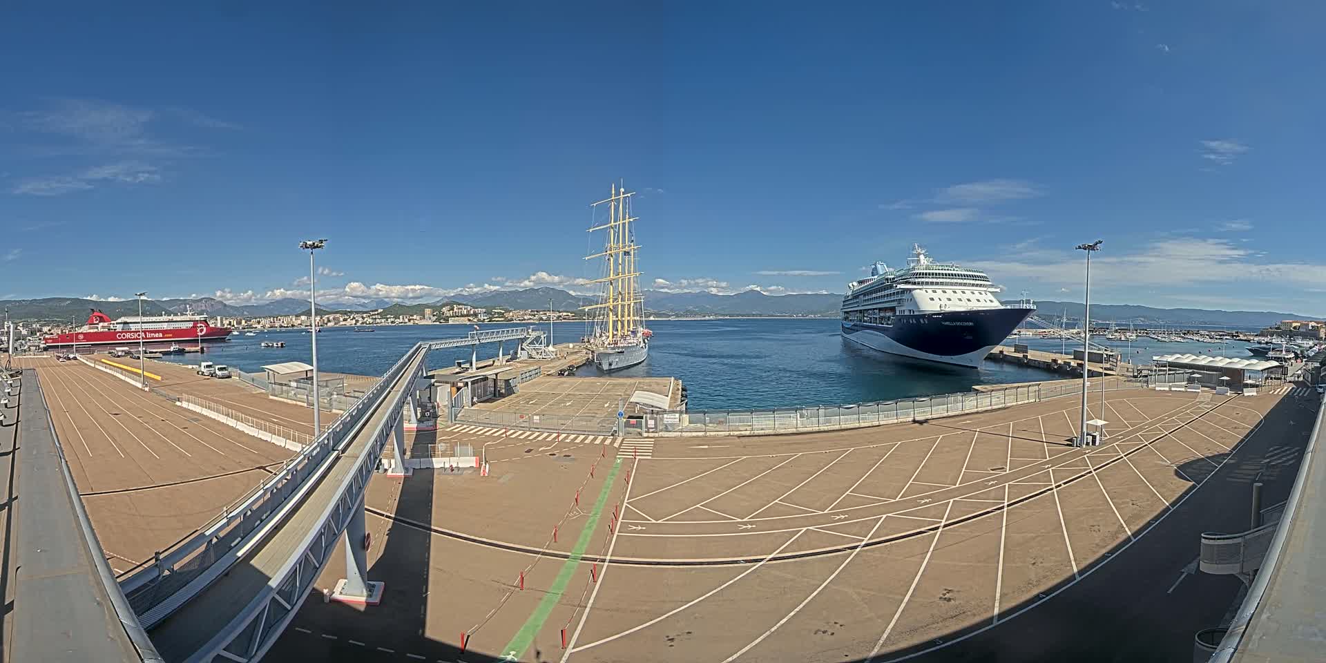 A cruise ship and a tall ship are docked at a port on a sunny day, with a ferry visible in the background.
