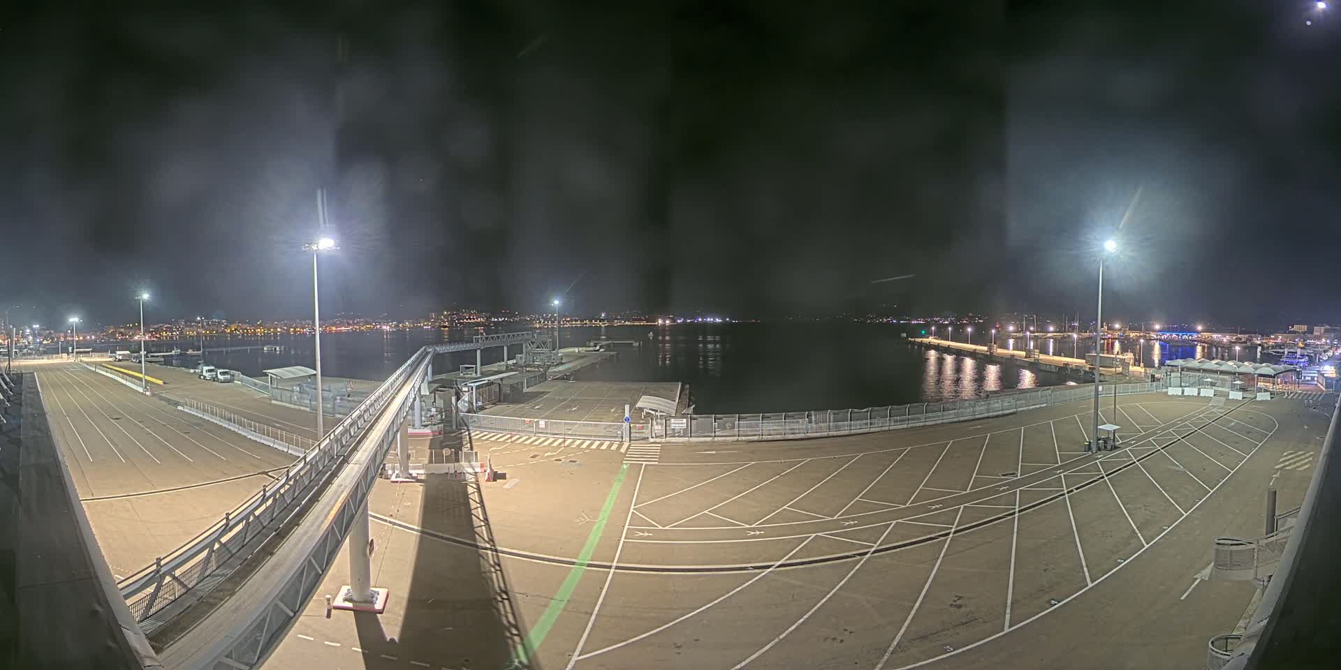 A nighttime view of an empty port area with a gangway, parking spaces, and a calm body of water reflecting city lights in the distance under a hazy sky.