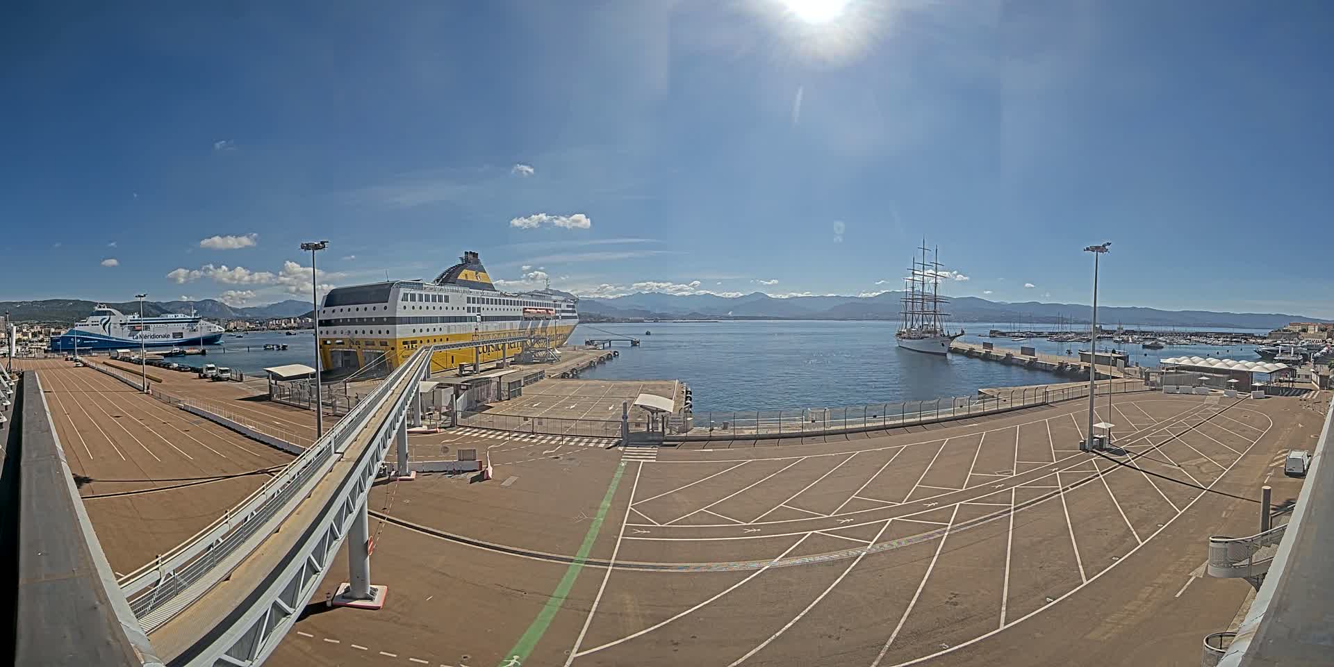 A sunny day at a port shows two large ships docked, one a ferry and the other a tall sailing ship, with a large empty parking lot and a walkway leading to the ferry.