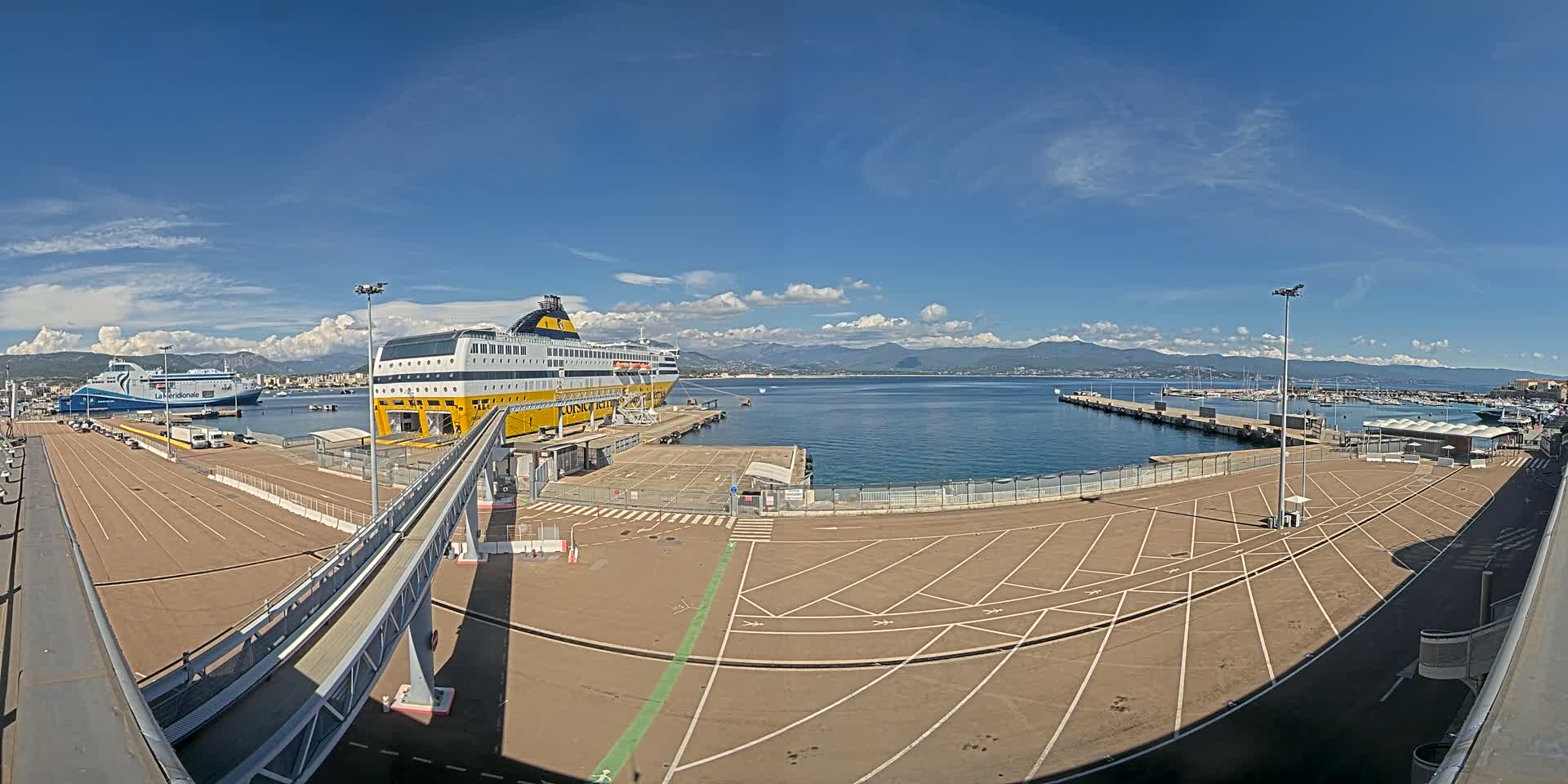 A large yellow and white ferry is docked at a port on a sunny day with a clear blue sky and mountains visible in the distance.