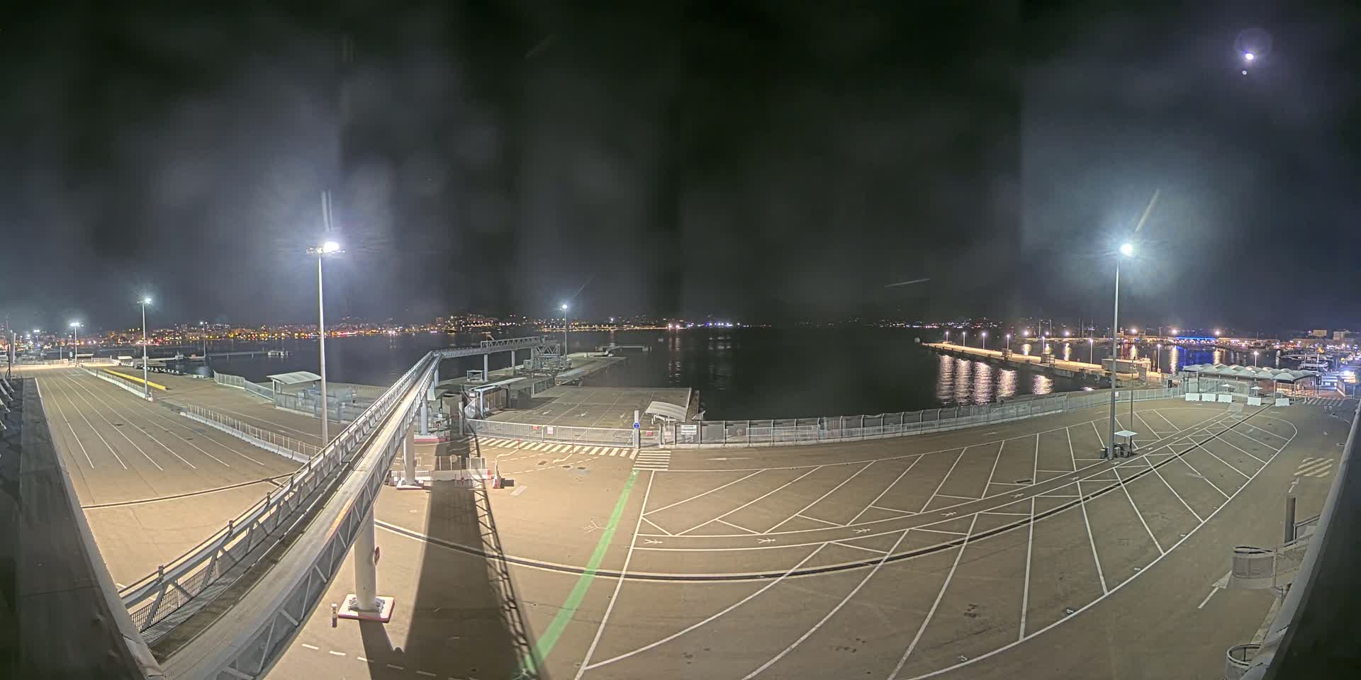 A nighttime view of a mostly empty port area with a walkway leading to a dock, illuminated by several bright lights, overlooking calm water and a distant city skyline under a slightly hazy sky.