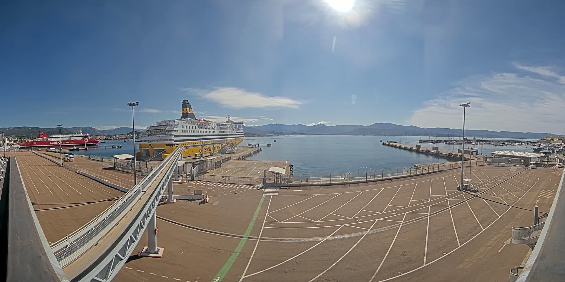 A large yellow and black ferry is docked at a port on a sunny day with calm water and mountains in the distance.