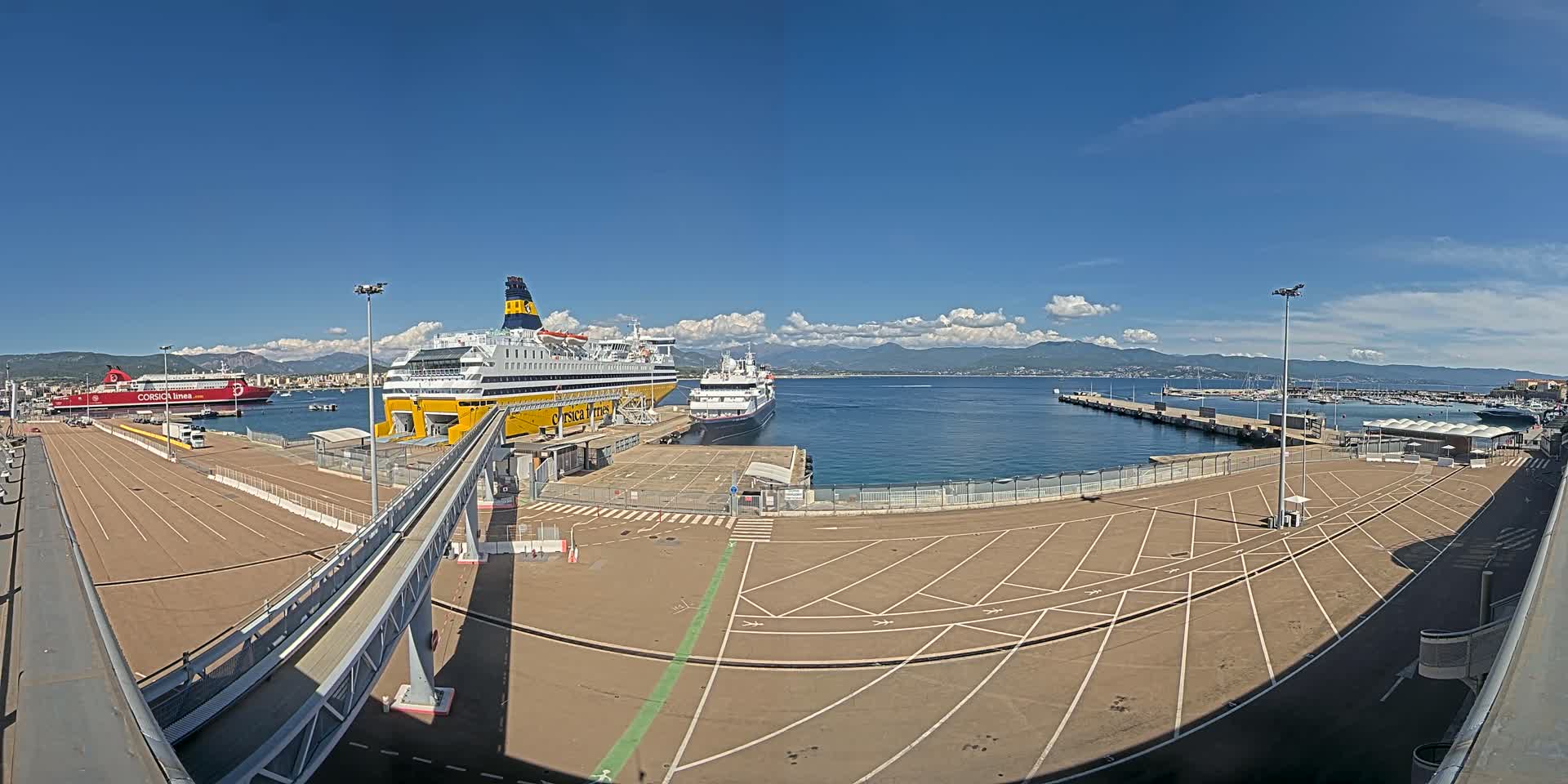 A large, mostly empty parking lot at a ferry terminal overlooks two docked ferries and a calm body of water under a mostly sunny sky.