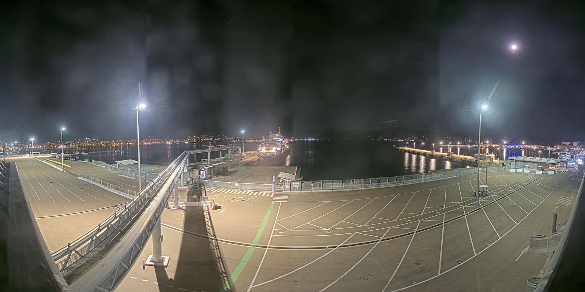 A nighttime view of an empty port area with a ship in the water, illuminated by several tall lights under a mostly clear sky with a visible moon.