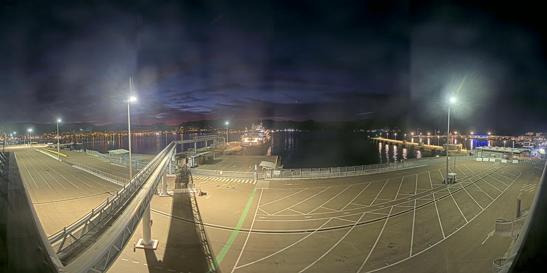 A nighttime view of a port with a ship docked, empty parking lots, and a gangway leading to it, under a mostly clear, dark sky.