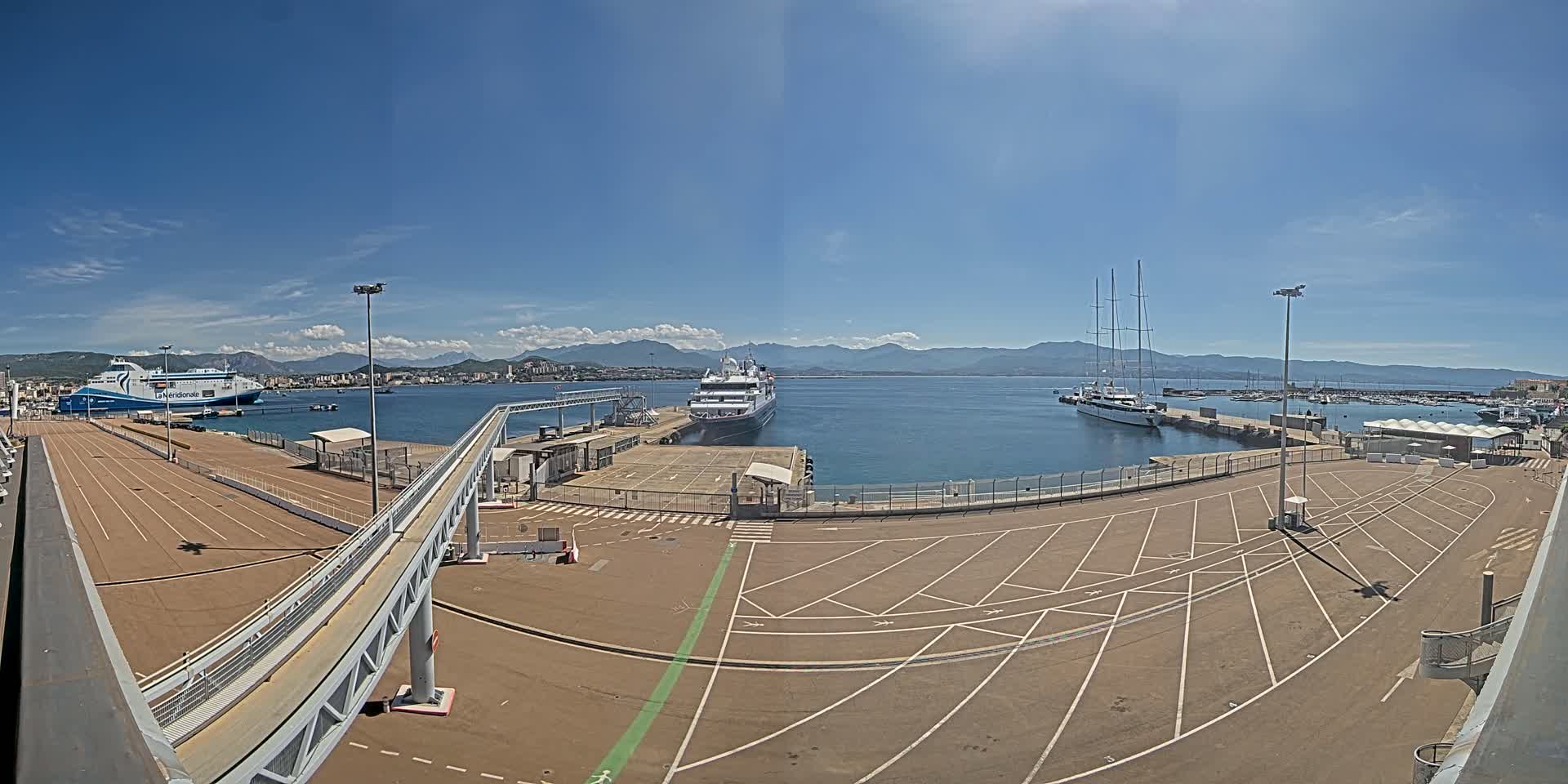 A sunny day at a port shows several ships docked, including a large ferry and a sailboat, with a large empty parking lot and a walkway leading to the docked ships.