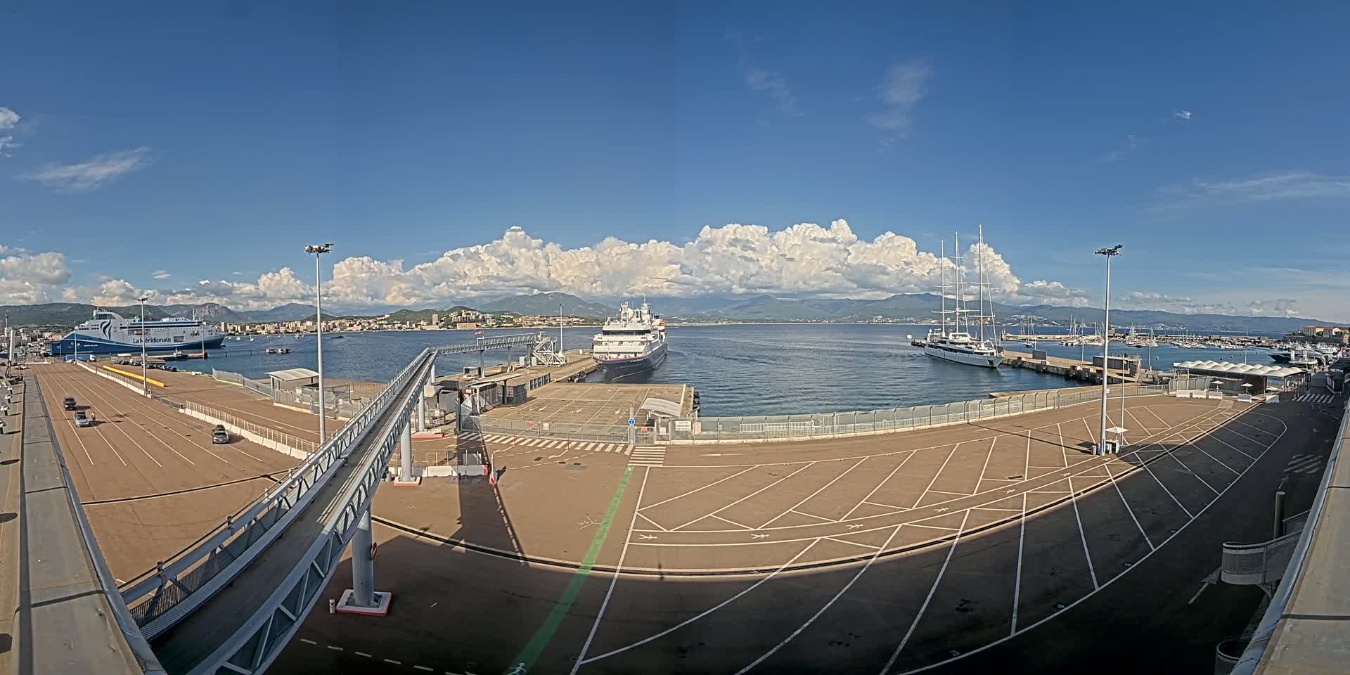 A cruise ship and ferry are docked at a port under a partly cloudy blue sky, with mountains visible in the distance.