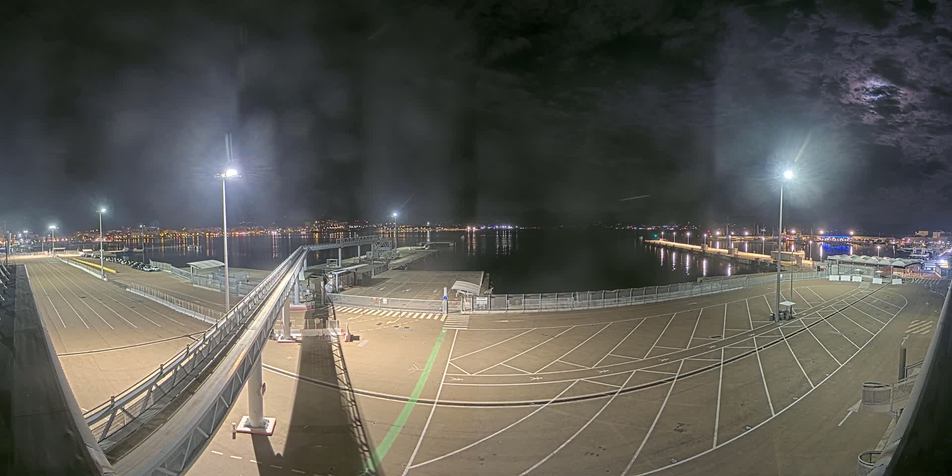 An empty, brightly lit nighttime port area with a gangway leading to a dock, overlooks a calm body of water and a city skyline.
