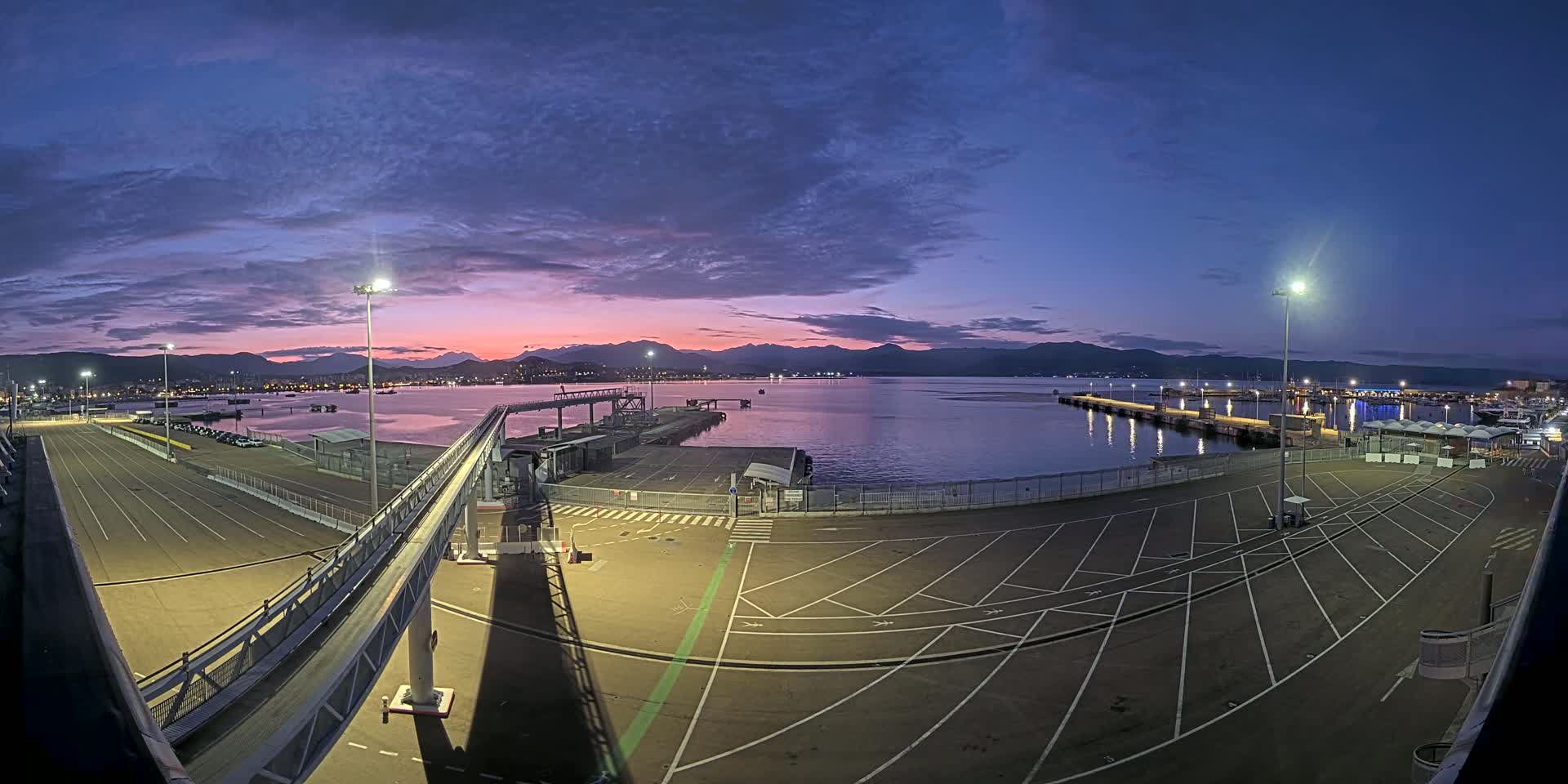 A mostly empty port parking lot at twilight, with a gangway leading to a dock, overlooks a calm body of water and mountains in the distance under a partly cloudy sky.