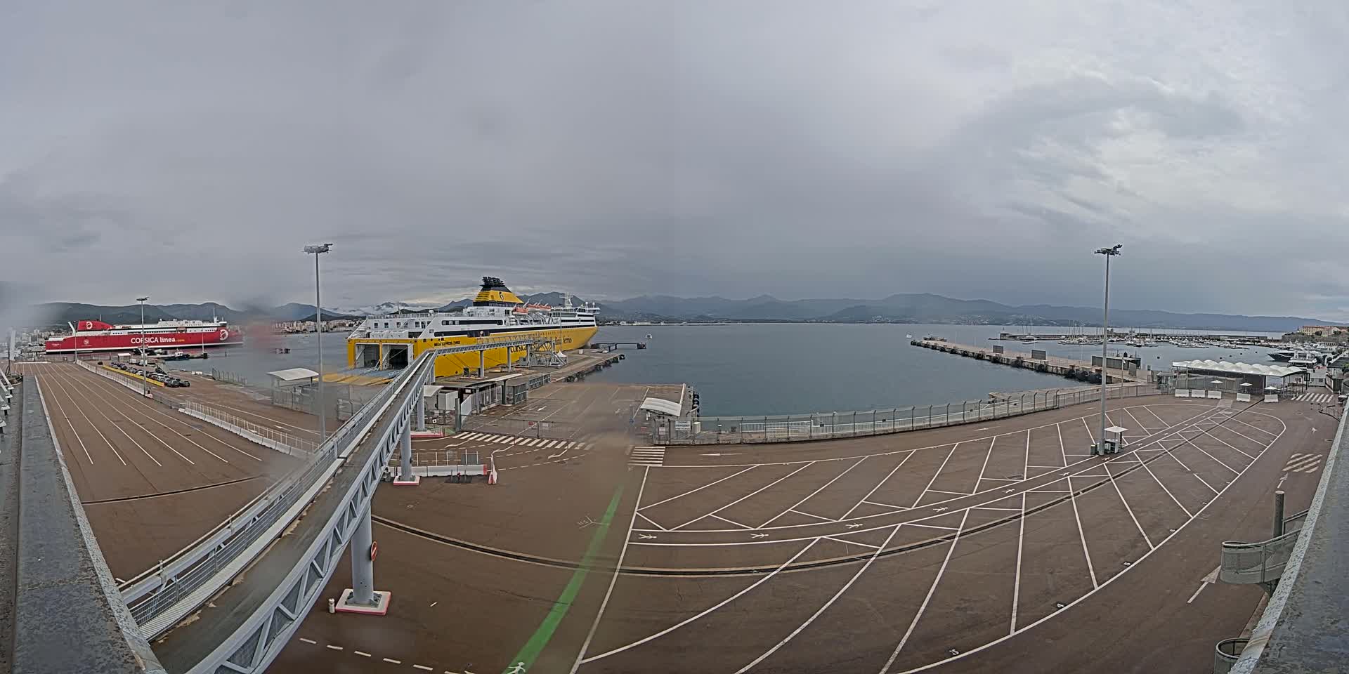 A ferry terminal on a cloudy day, showing two docked ferries, a large empty parking lot, and a walkway leading to one of the ships.