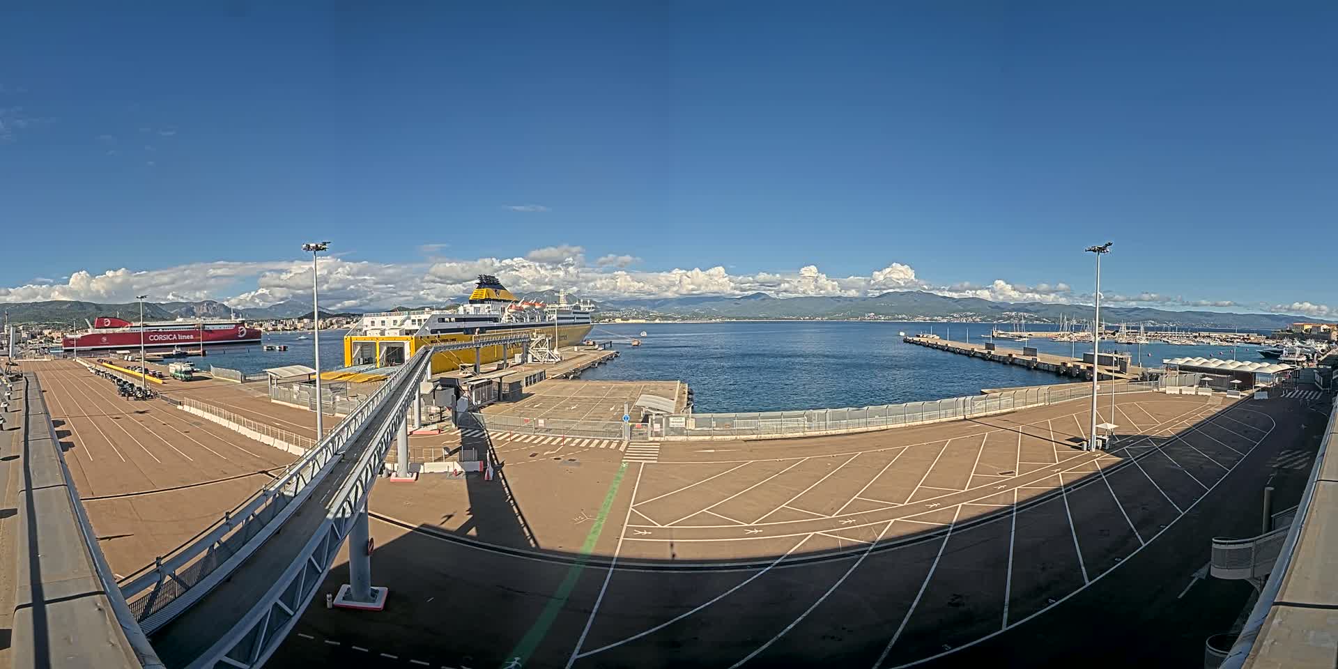 A ferry terminal with two large ferries docked, under a mostly sunny sky with some clouds, overlooks a calm body of water with mountains in the distance.