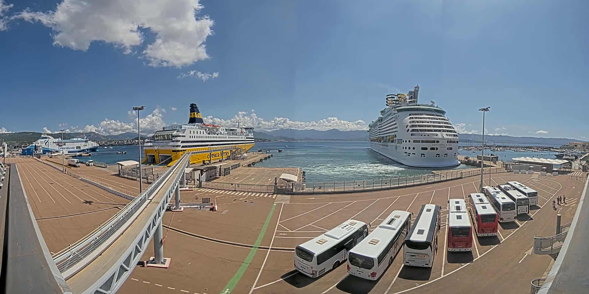 A sunny day at a port shows two large passenger ships docked, one a ferry and the other a cruise ship, with several buses parked nearby.
