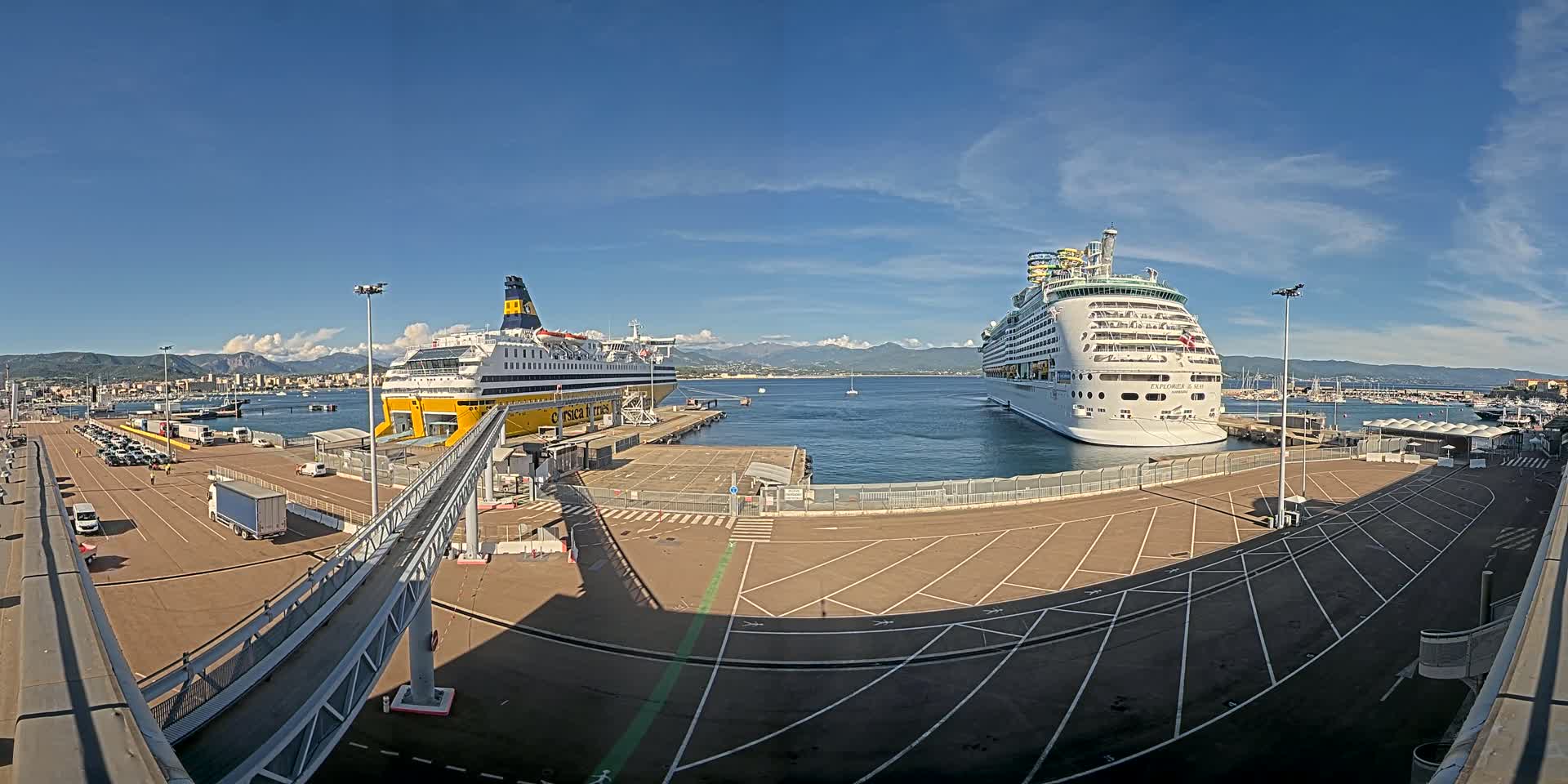 A large ferry and a cruise ship are docked at a port on a sunny day with mountains in the background.