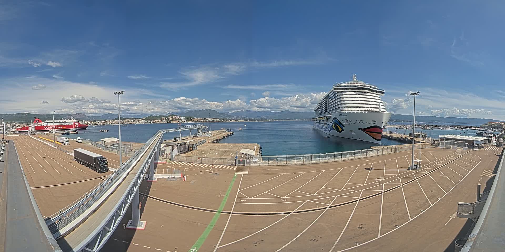 A large cruise ship is docked at a port on a sunny day with partly cloudy skies, alongside a ferry and numerous parked vehicles.