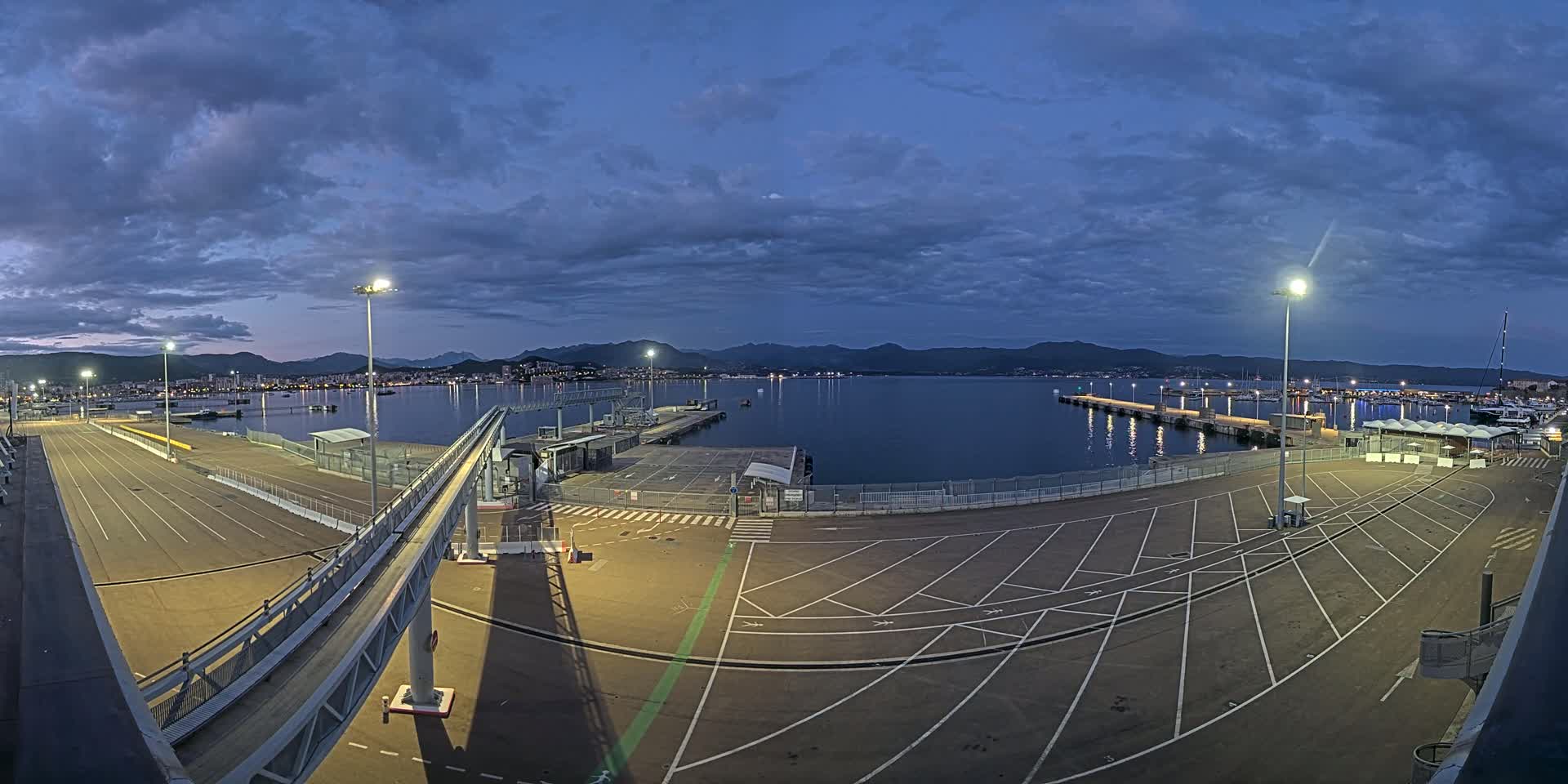 An empty, illuminated parking lot at a harbor overlooks a calm body of water under a twilight sky with cloudy conditions.