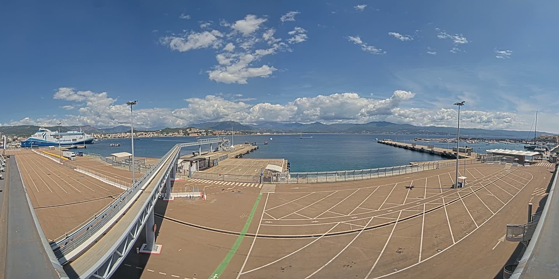 A ferry is docked at a port under a partly cloudy blue sky, with mountains visible in the distance.